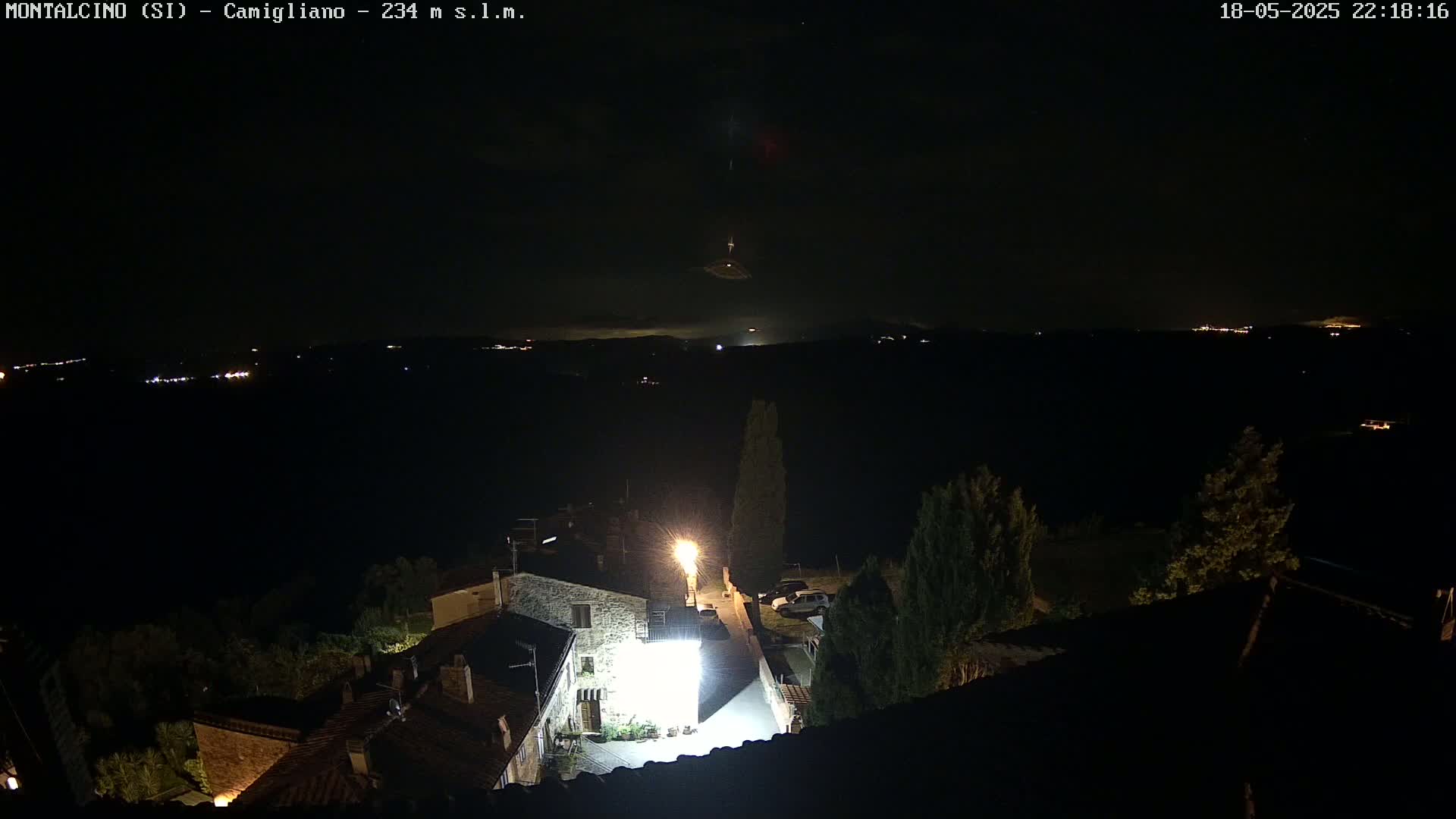 A nighttime view of a small village illuminated by streetlights, overlooking a dark valley with distant lights under a mostly clear sky.