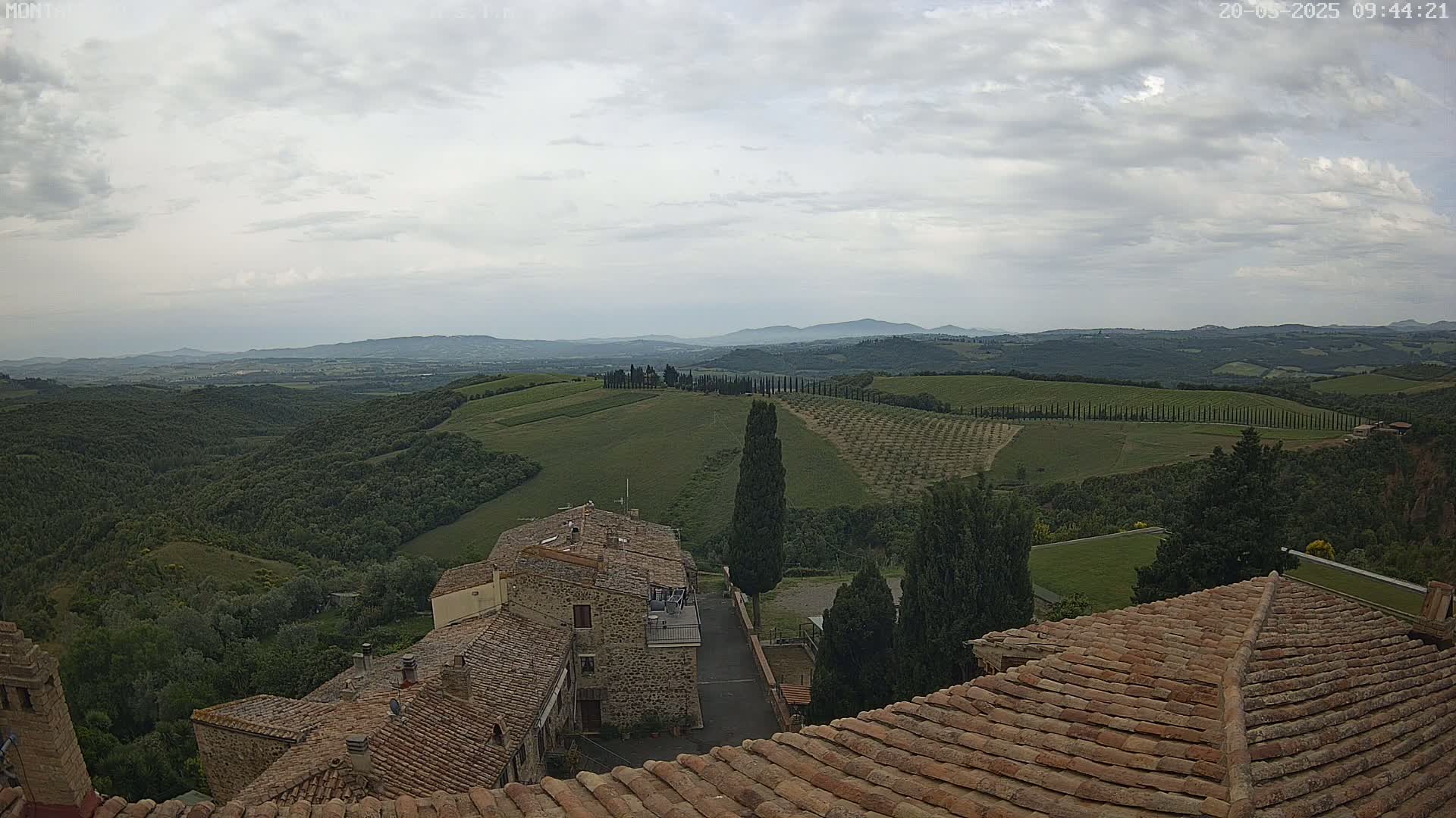 Overlooking terracotta rooftops of buildings, a sprawling landscape of rolling green hills, rows of trees, and distant mountains under a cloudy sky.