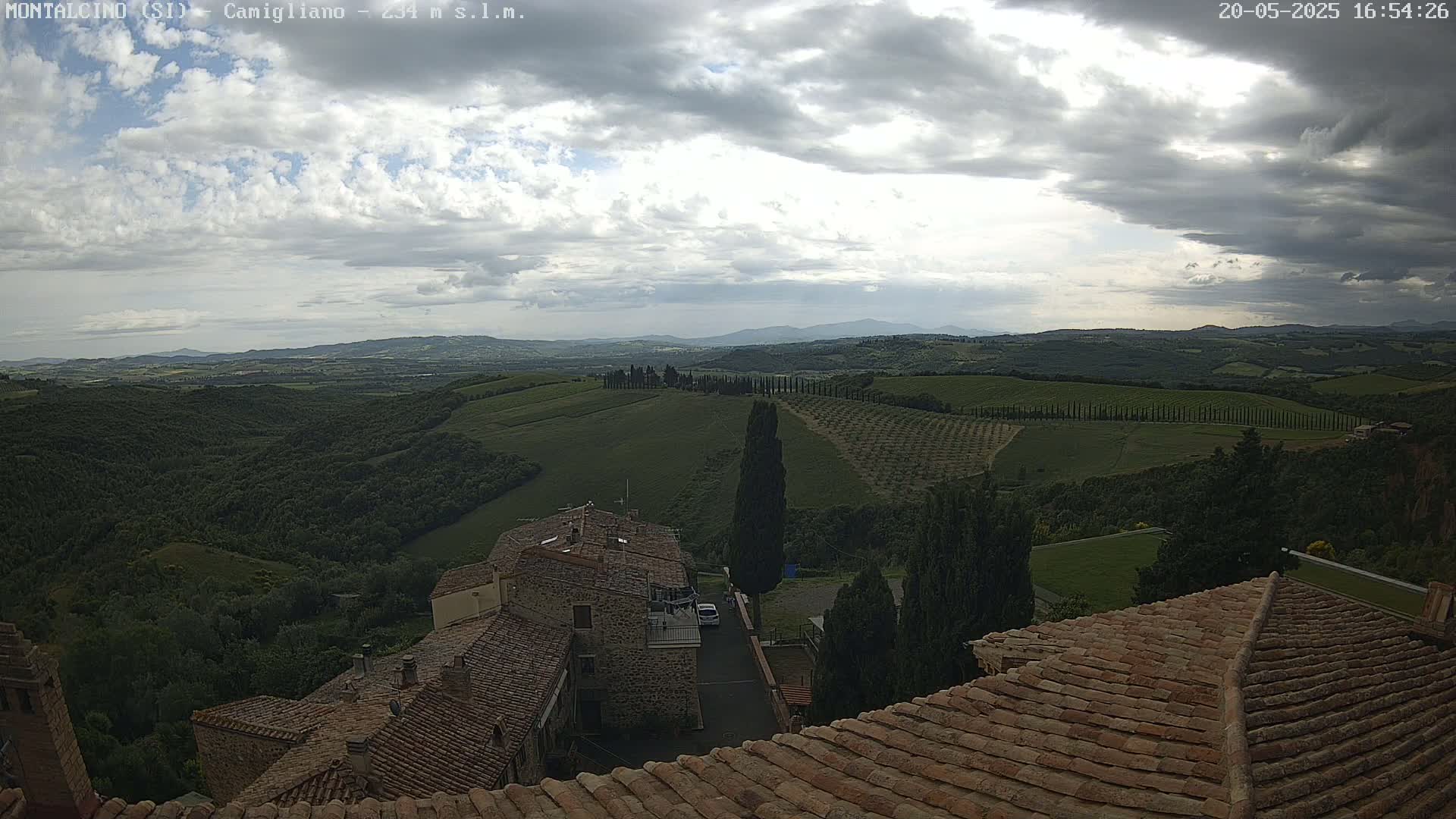 A view from a rooftop shows rolling hills of olive groves and vineyards under a partly cloudy sky.