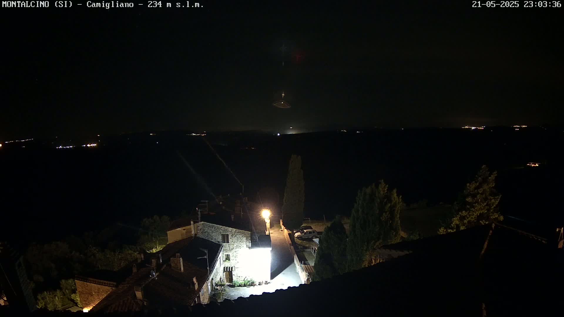 A nighttime aerial view shows a small village illuminated by a streetlight, with a dark, clear sky above and distant lights visible on the horizon.