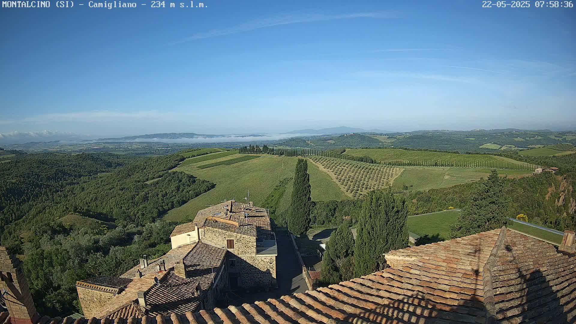 A high-angle view shows a clear, sunny day overlooking a picturesque Tuscan landscape of rolling hills, vineyards, and olive groves, with stone buildings and terracotta rooftops in the foreground.