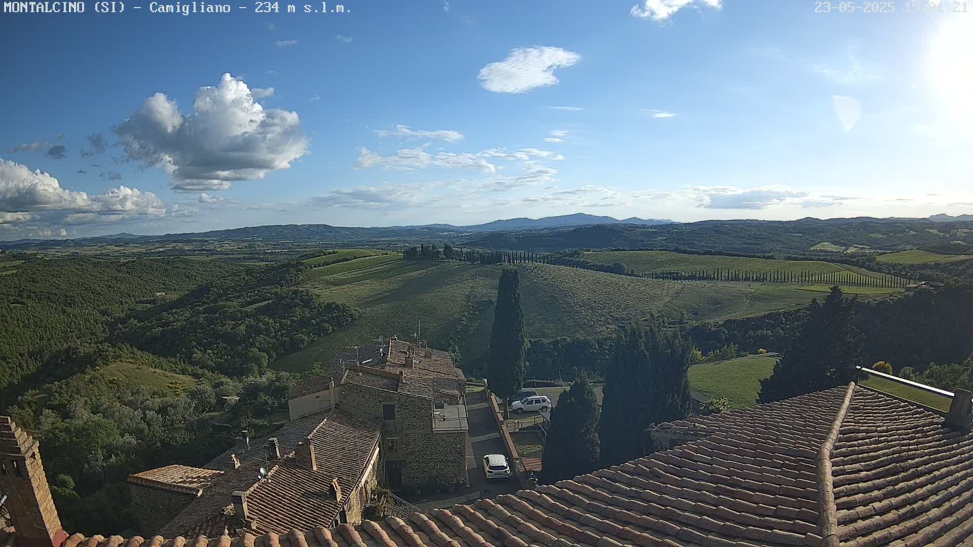 A partly cloudy sky overlooks a hilly, green landscape dotted with buildings and rows of trees, viewed from the terracotta rooftops of a village.