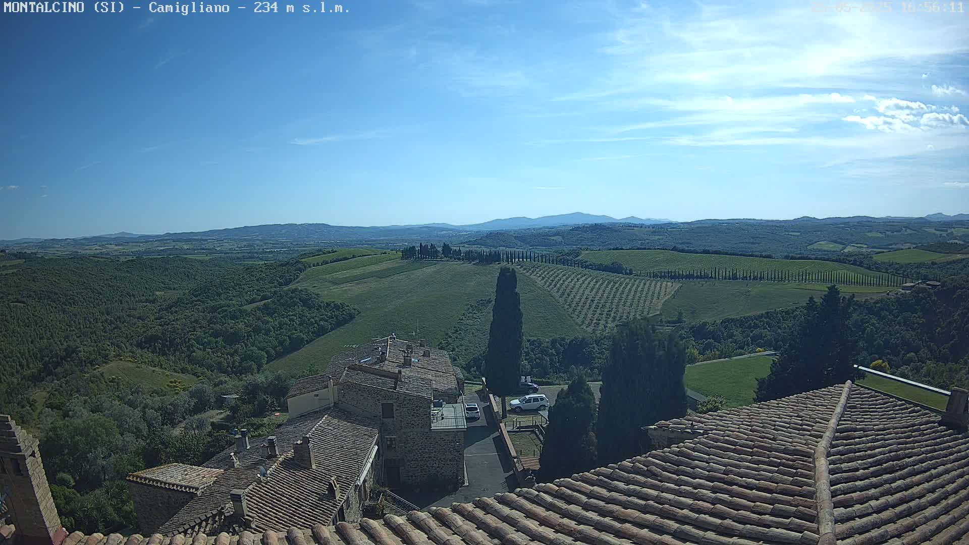 A view from a tiled rooftop shows a sunny, hilly landscape with rows of trees and buildings, under a mostly clear blue sky.