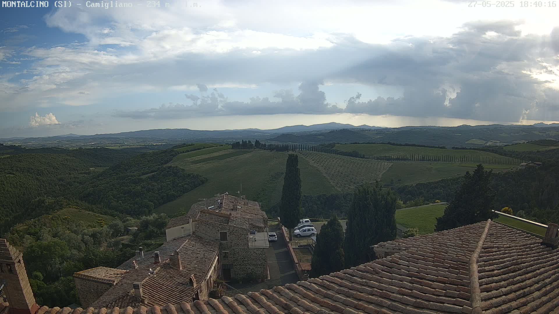 Overlooking a hilly landscape of green fields and cypress trees under a partly cloudy sky, stone rooftops of buildings are in the foreground.