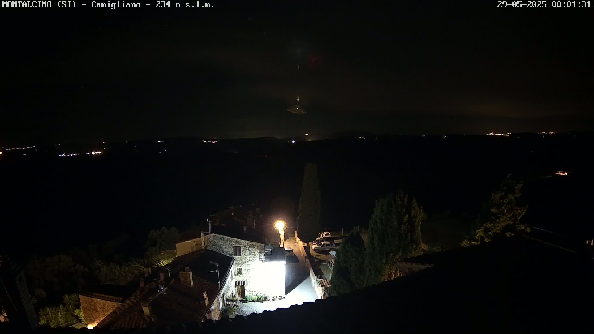 A nighttime, overhead view of a small village with dimly lit buildings and a few cars, overlooking a dark landscape speckled with distant lights under a mostly clear sky.