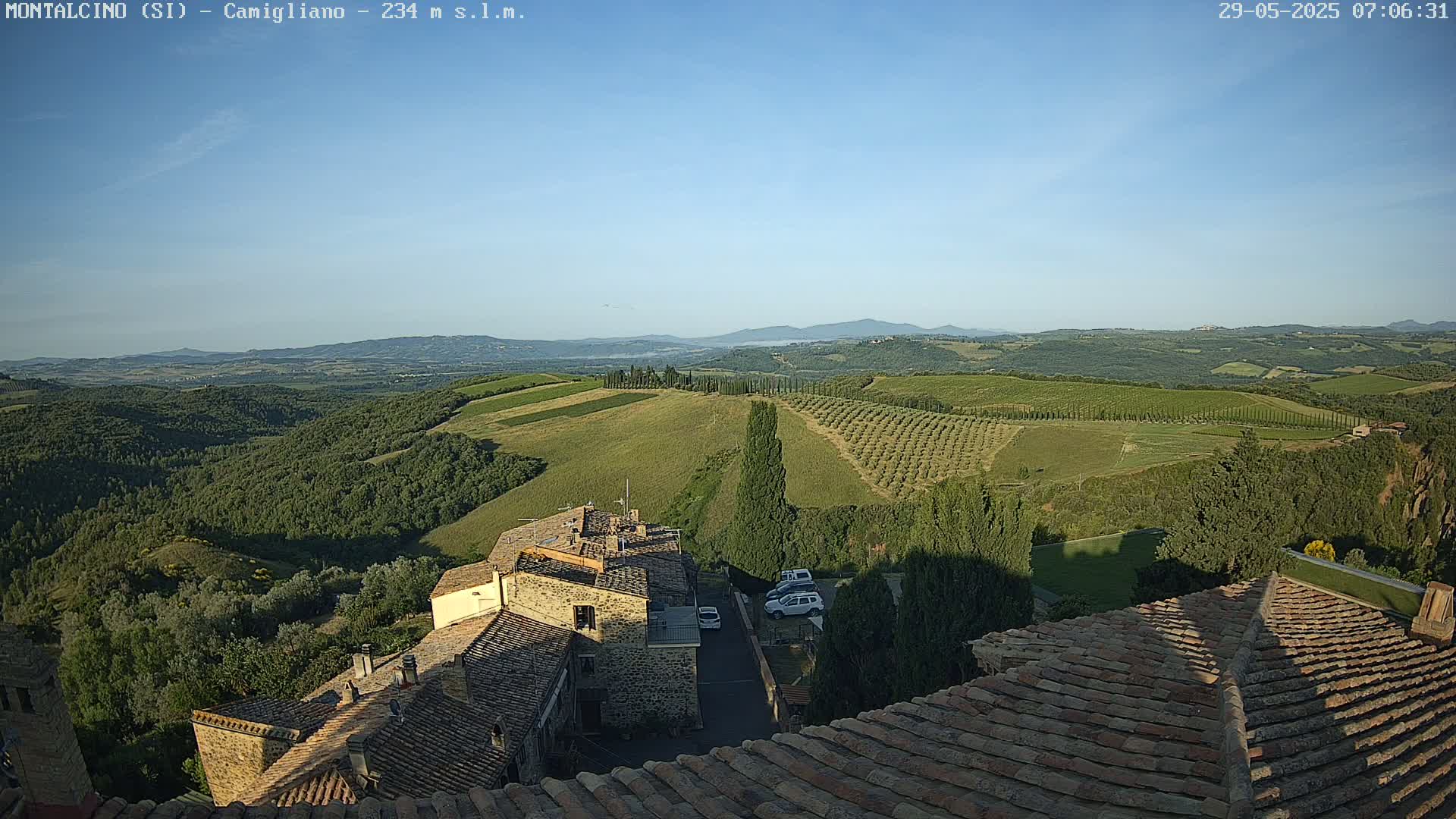 A view from a rooftop shows a clear, sunny day over a rolling landscape of green hills, vineyards, and olive groves, with a small village nestled below.