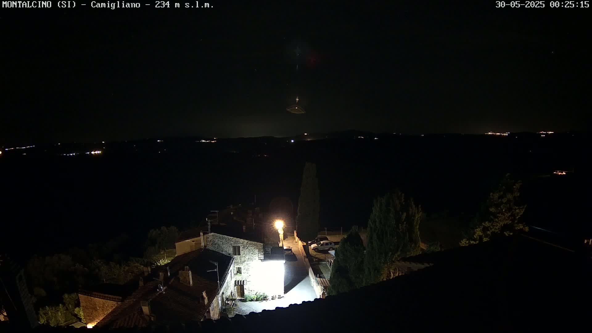 A nighttime view of a small village illuminated by a few lights, with a dark, clear sky showing distant lights and what appears to be a meteor or other celestial event.