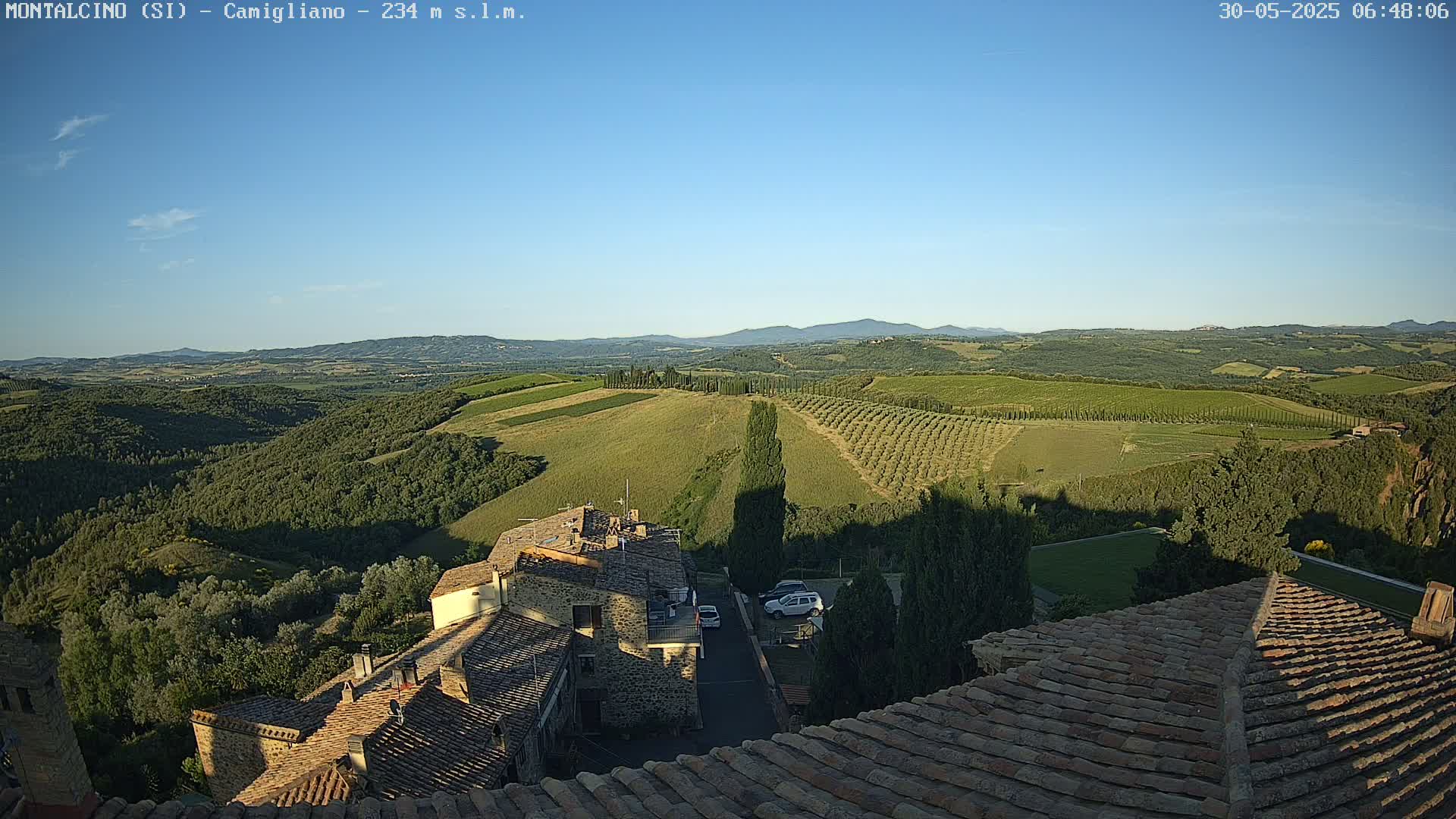 A clear, sunny view from a rooftop shows rolling hills of farmland and olive groves, with a small village nestled below.