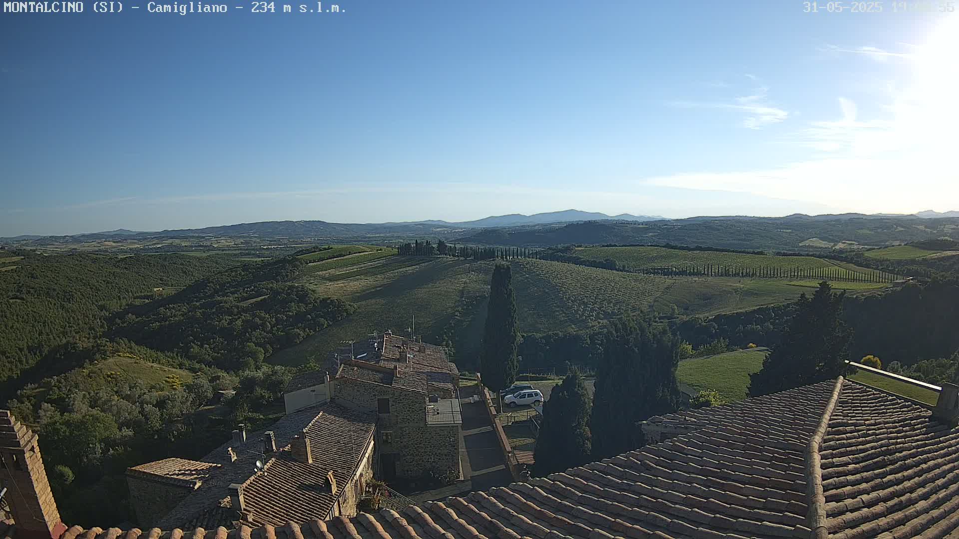 A view from a rooftop shows a clear, sunny day over rolling green hills dotted with trees and buildings, extending to distant mountains under a mostly clear blue sky.