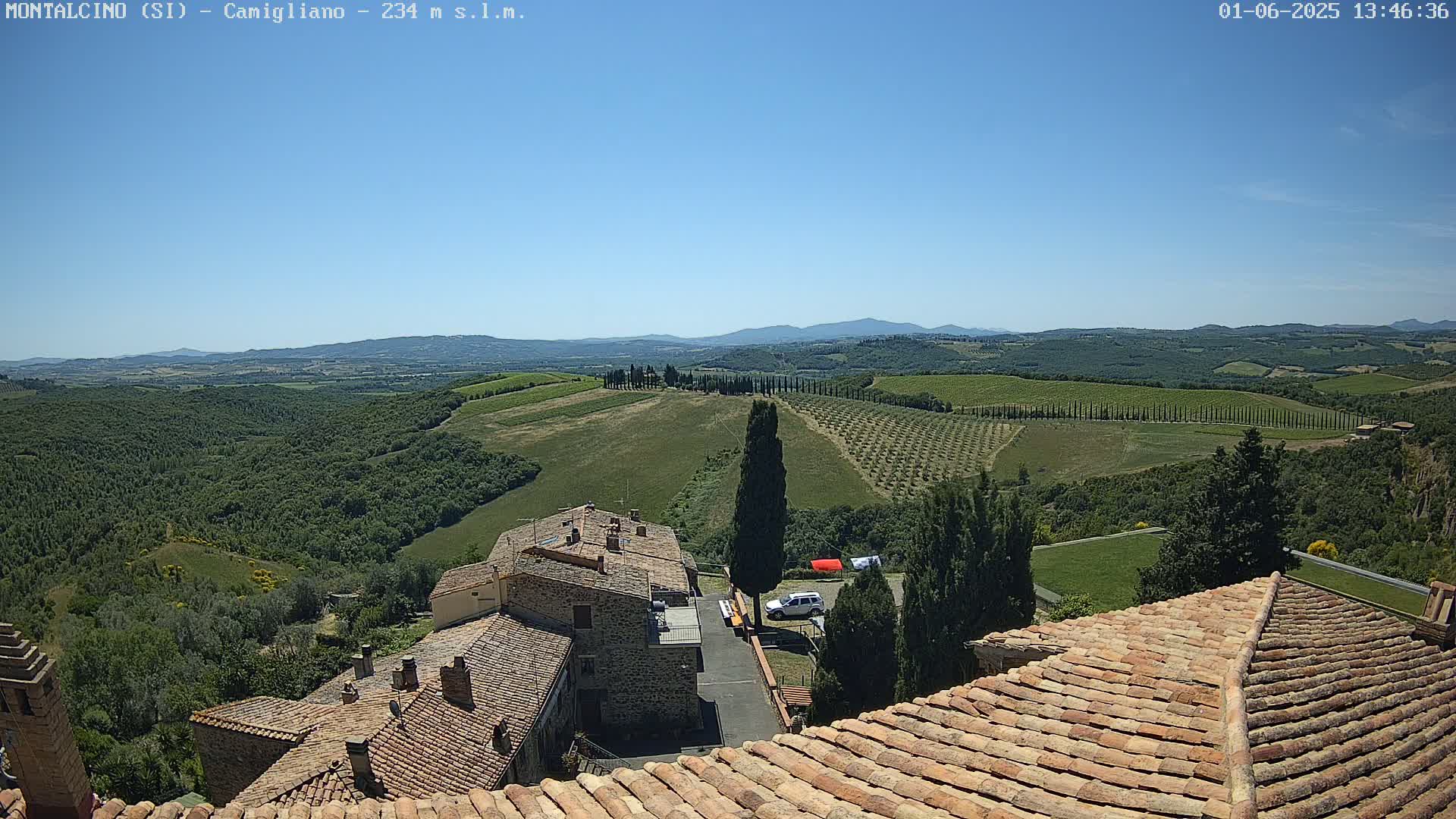 A sprawling vista of rolling green hills dotted with trees and vineyards under a clear blue sky, overlooks the terracotta roofs of stone buildings.