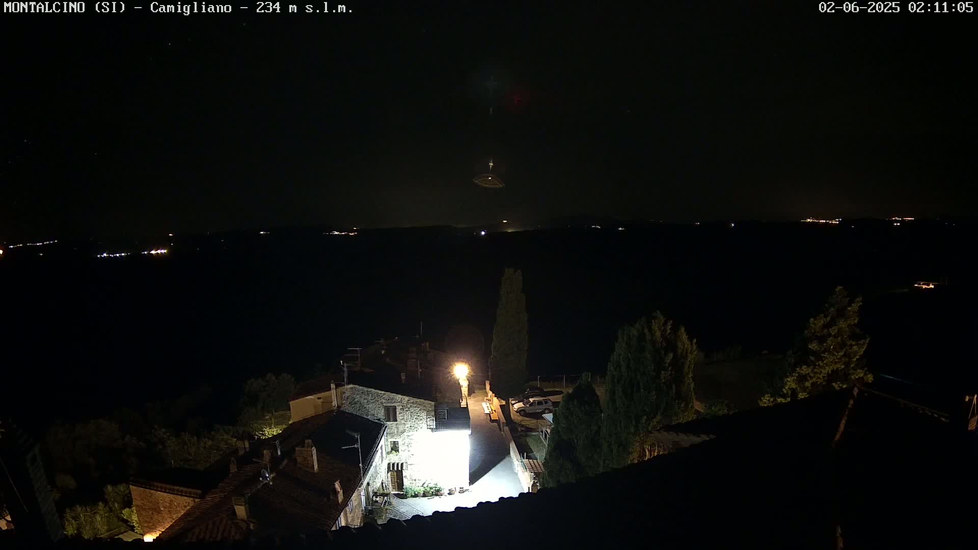 A nighttime aerial view of a small village illuminated by streetlights, overlooking a dark landscape dotted with distant lights under a clear, starry sky.