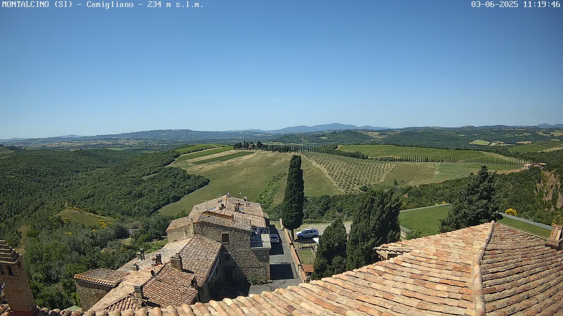 A view from a terracotta roof shows a sunny, clear-day vista of rolling hills dotted with vineyards and olive groves, overlooking stone buildings in a village.