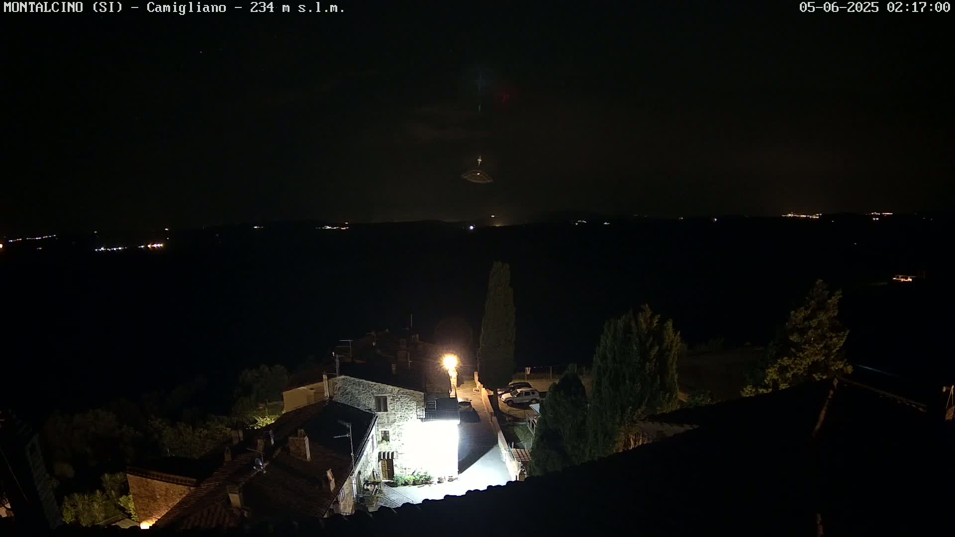 A nighttime view of a small town illuminated by streetlights, with a distant horizon showing faint lights and a partly cloudy sky.