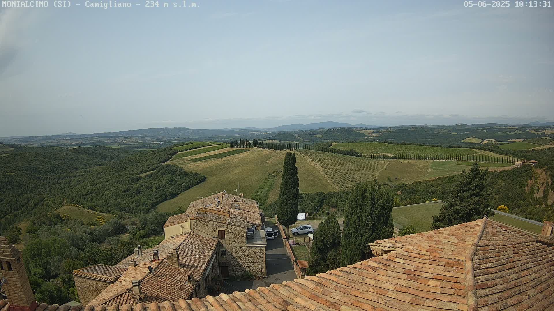 A view from a tiled rooftop shows a village nestled amongst rolling green hills dotted with trees and vineyards under a clear sky.