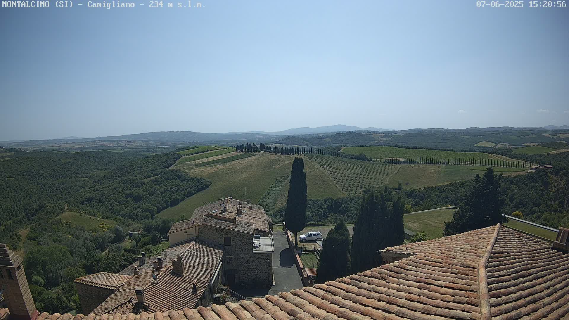 A clear, sunny day reveals a Tuscan landscape of rolling hills dotted with vineyards and cypress trees, overlooking a cluster of stone buildings with terracotta roofs.