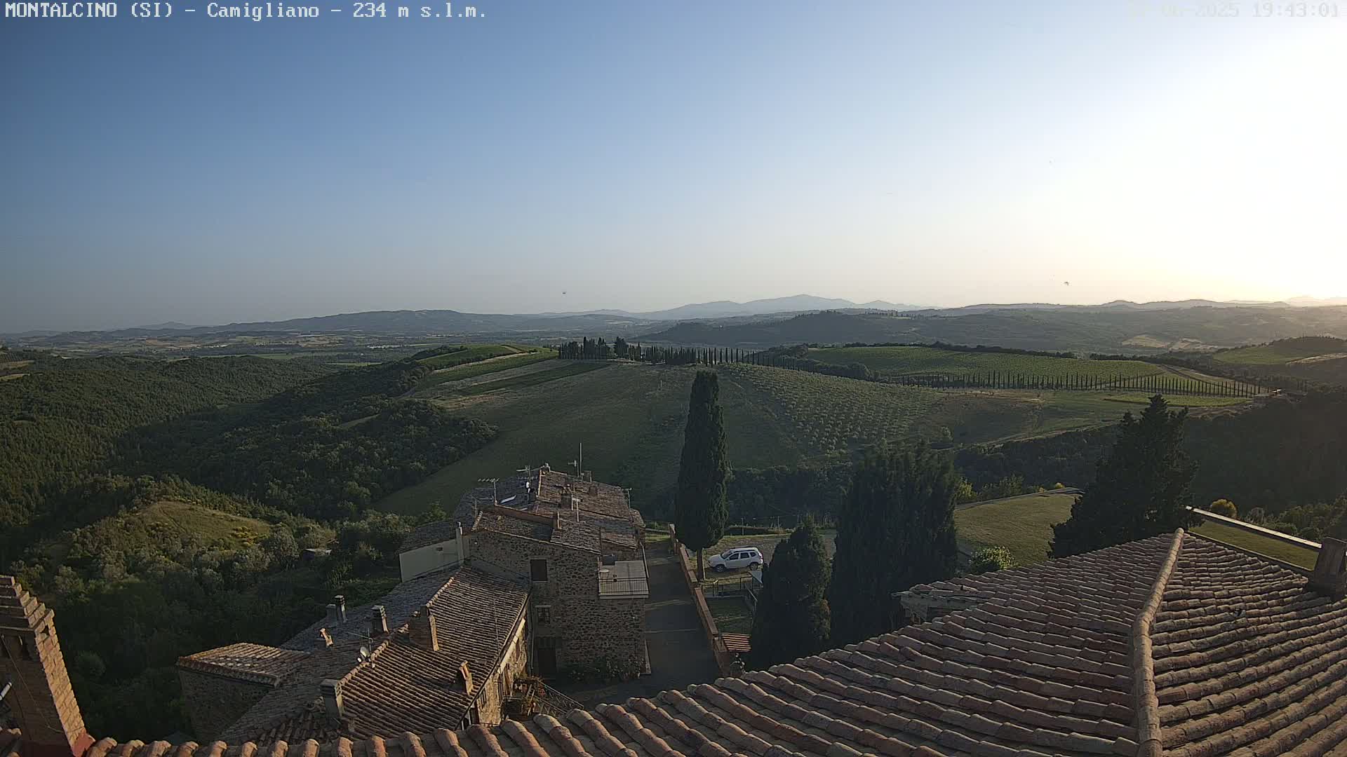 A view from a rooftop shows rolling hills dotted with olive trees and vineyards under a clear, bright sky.
