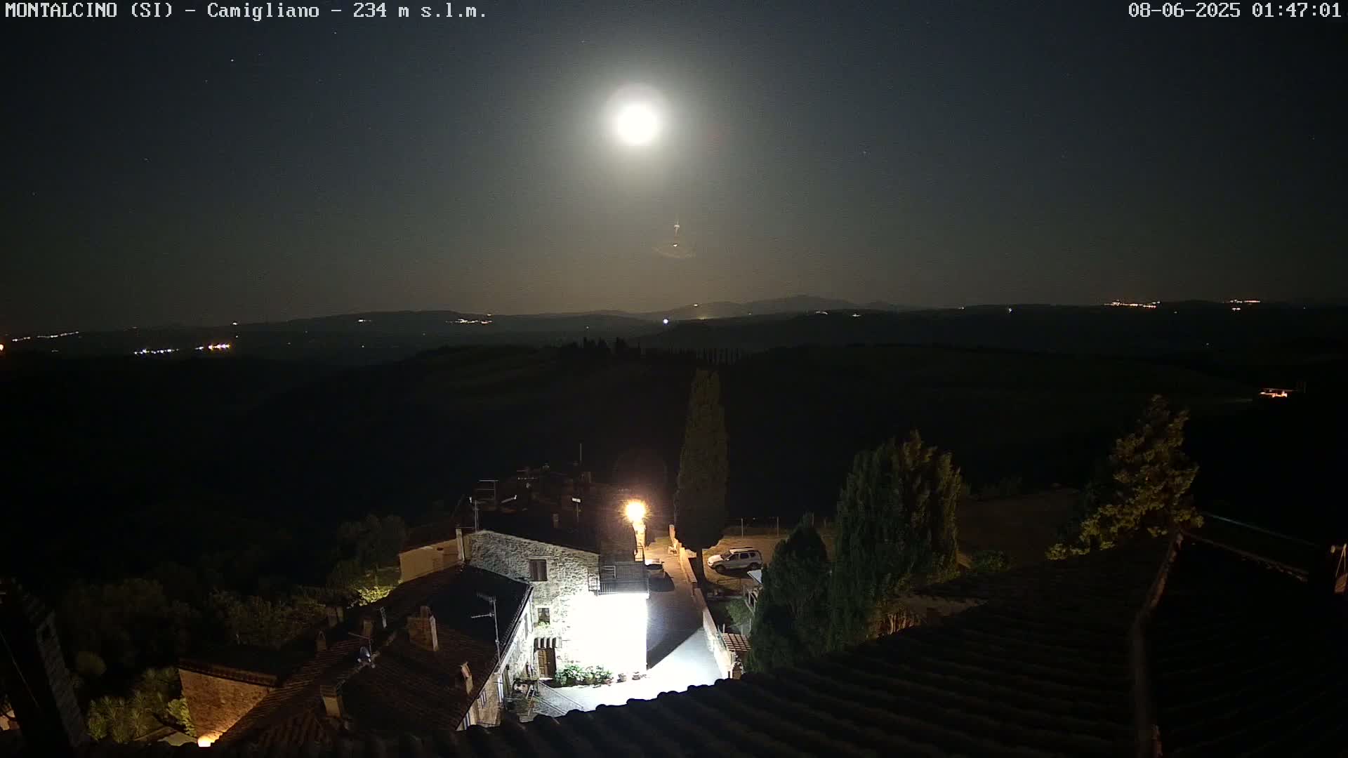 A brightly lit, moonlit village nestled in a dark, rolling landscape under a clear night sky.