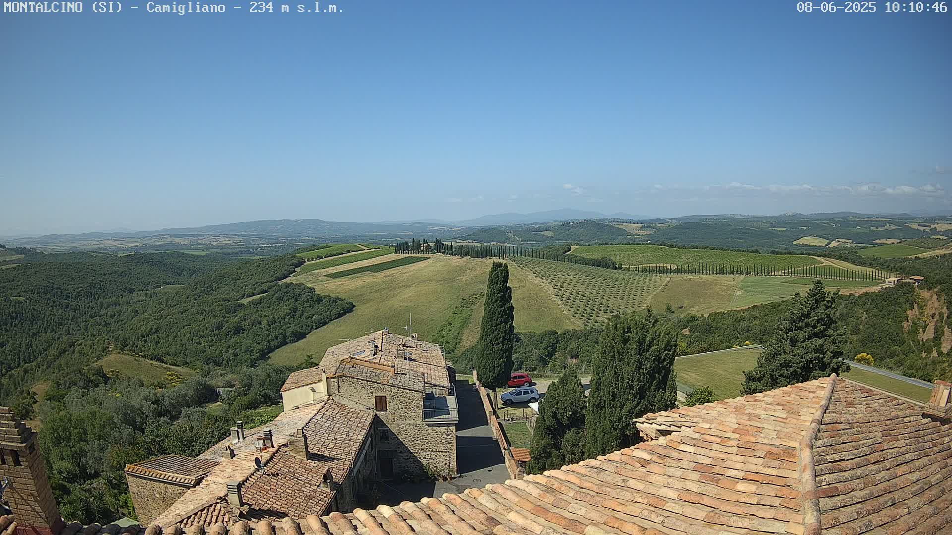 Under a clear blue sky, a view from a rooftop shows terracotta-tiled buildings nestled amongst rolling hills dotted with olive groves and vineyards, extending to a distant mountain range.
