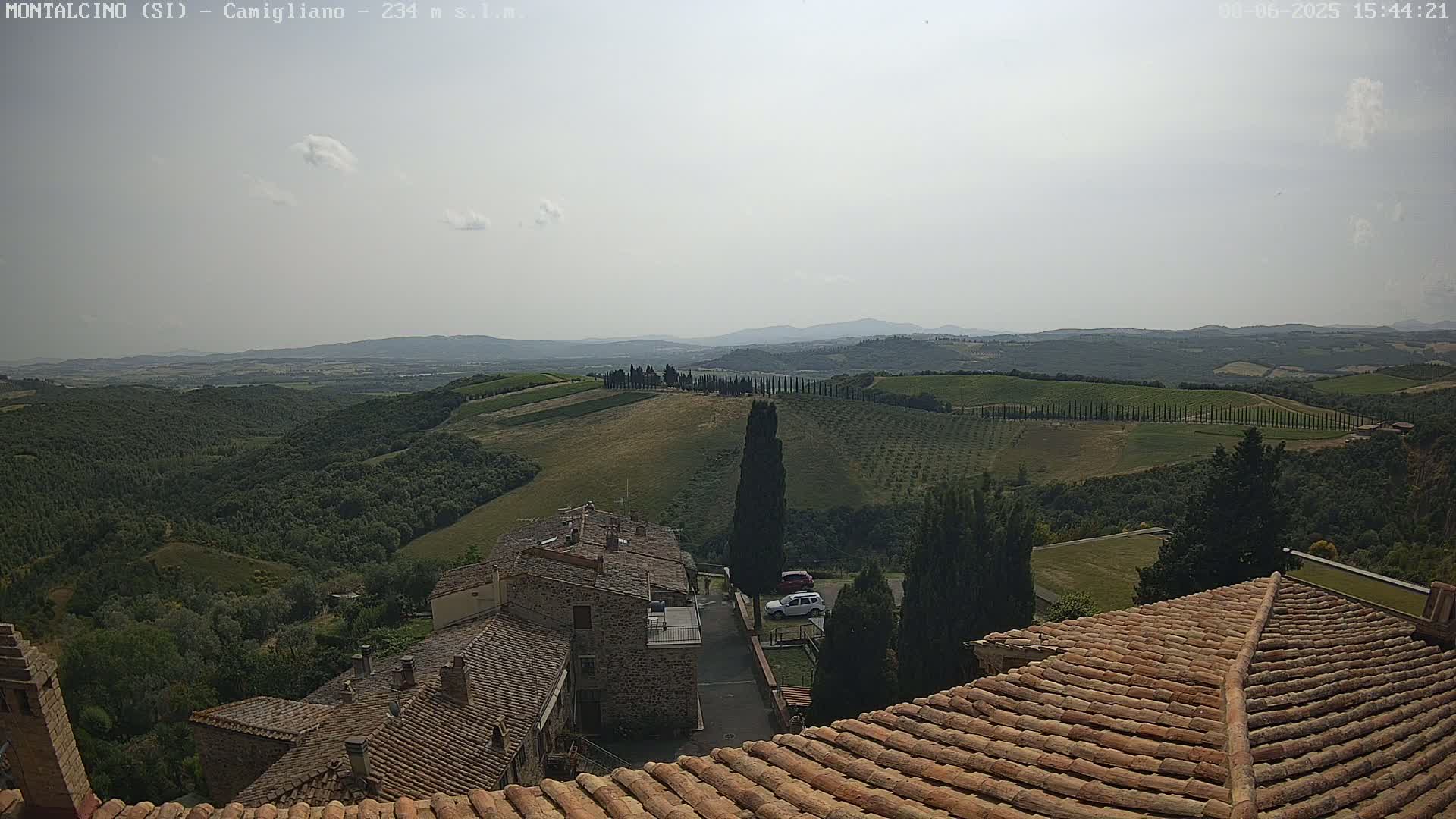 A view from a terracotta roof shows a hazy, sunny day over rolling hills of vineyards and olive groves, with a small village nestled below.