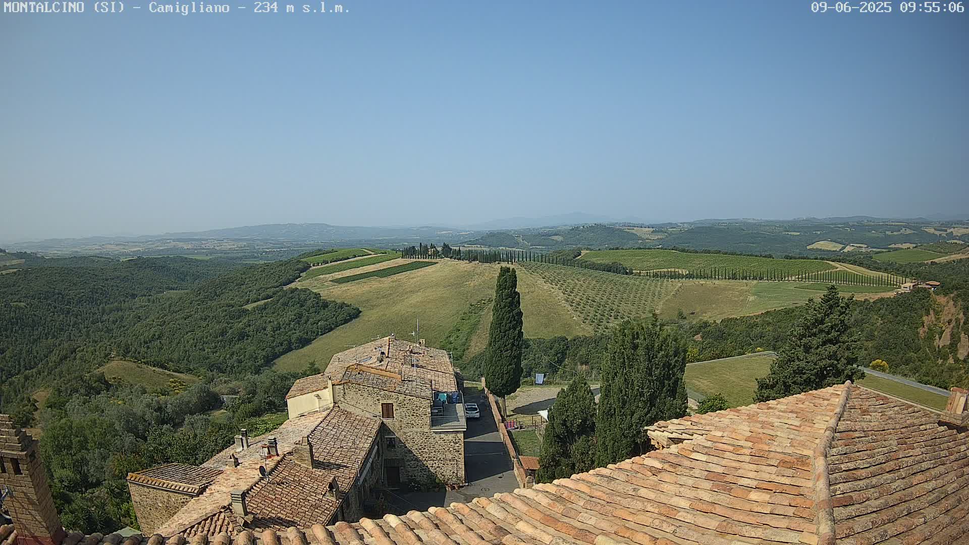 A clear, sunny day reveals a Tuscan landscape of rolling hills dotted with olive groves and vineyards, viewed from atop a village of terracotta-roofed buildings.