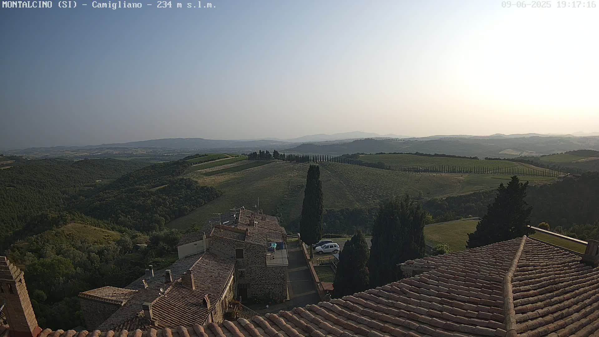 A view from a rooftop shows rolling hills of vineyards and olive groves under a clear, hazy sky.