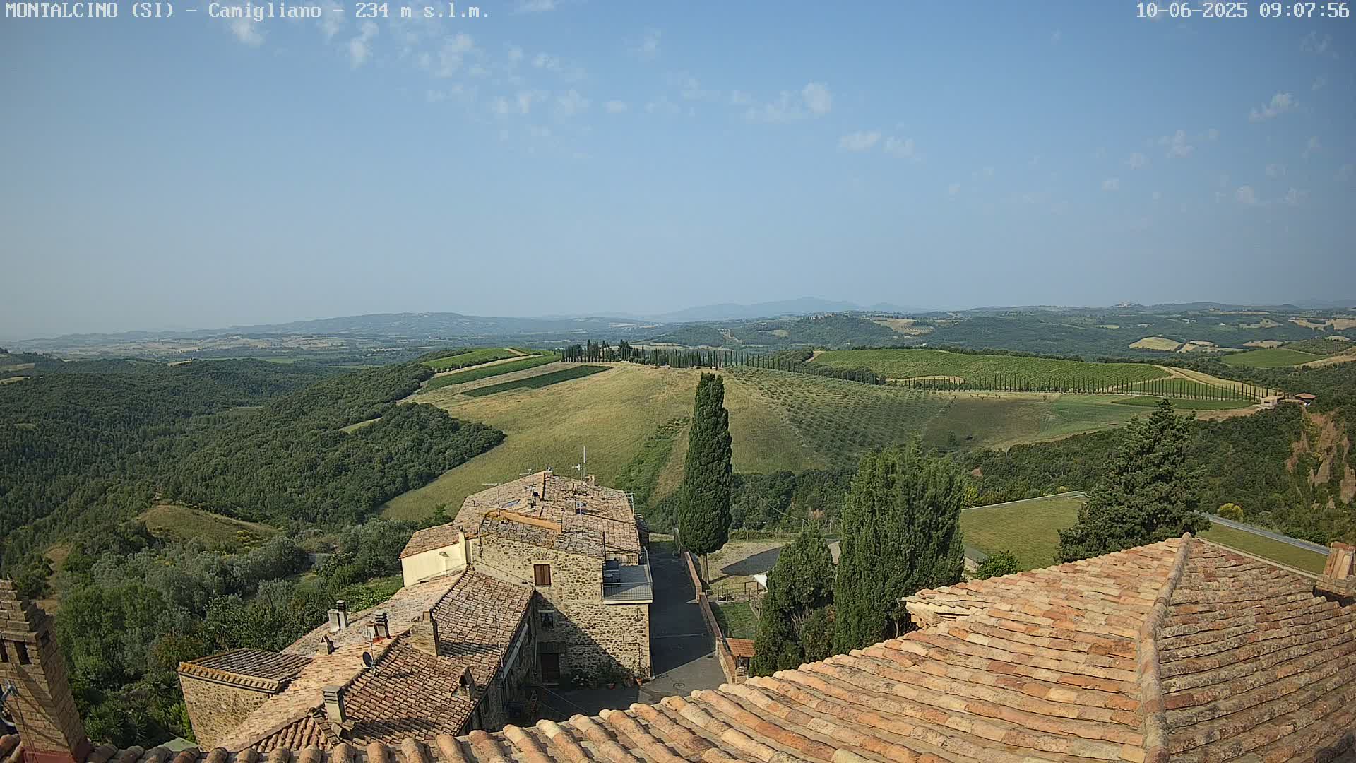 A high-angle view on a clear day shows a sun-drenched Tuscan landscape of rolling hills dotted with vineyards, olive groves, and cypress trees, overlooking terracotta-roofed buildings.