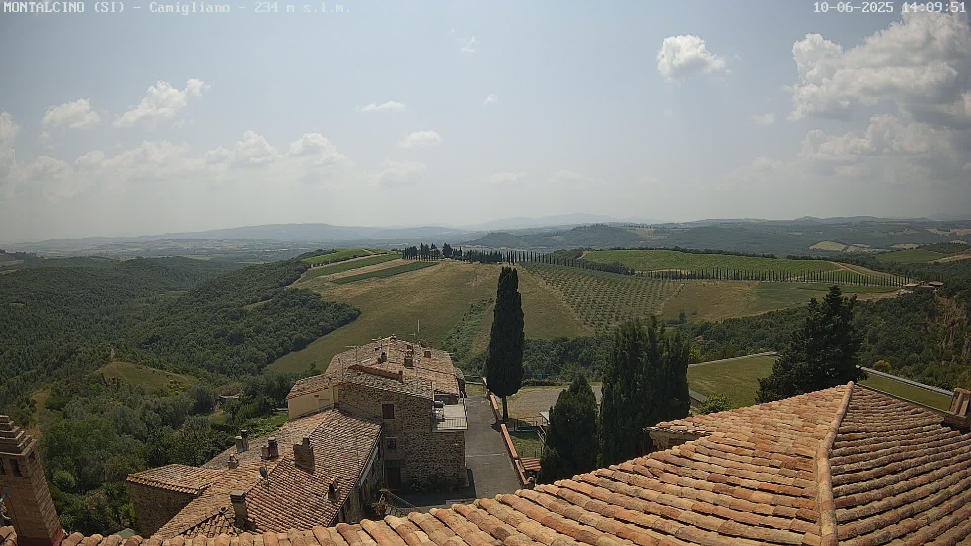 A partly cloudy sky overlooks a hilly landscape of olive groves and vineyards, with a view of terracotta rooftops and stone buildings in the foreground.