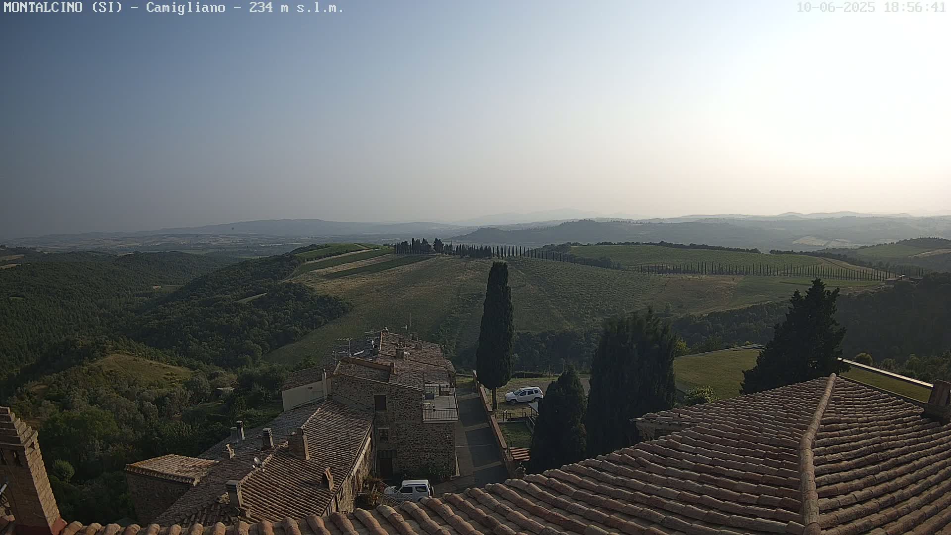 A hazy, sunny view from a rooftop shows rolling hills covered in green vegetation and rows of cypress trees, overlooking a small village with terracotta-tiled roofs.
