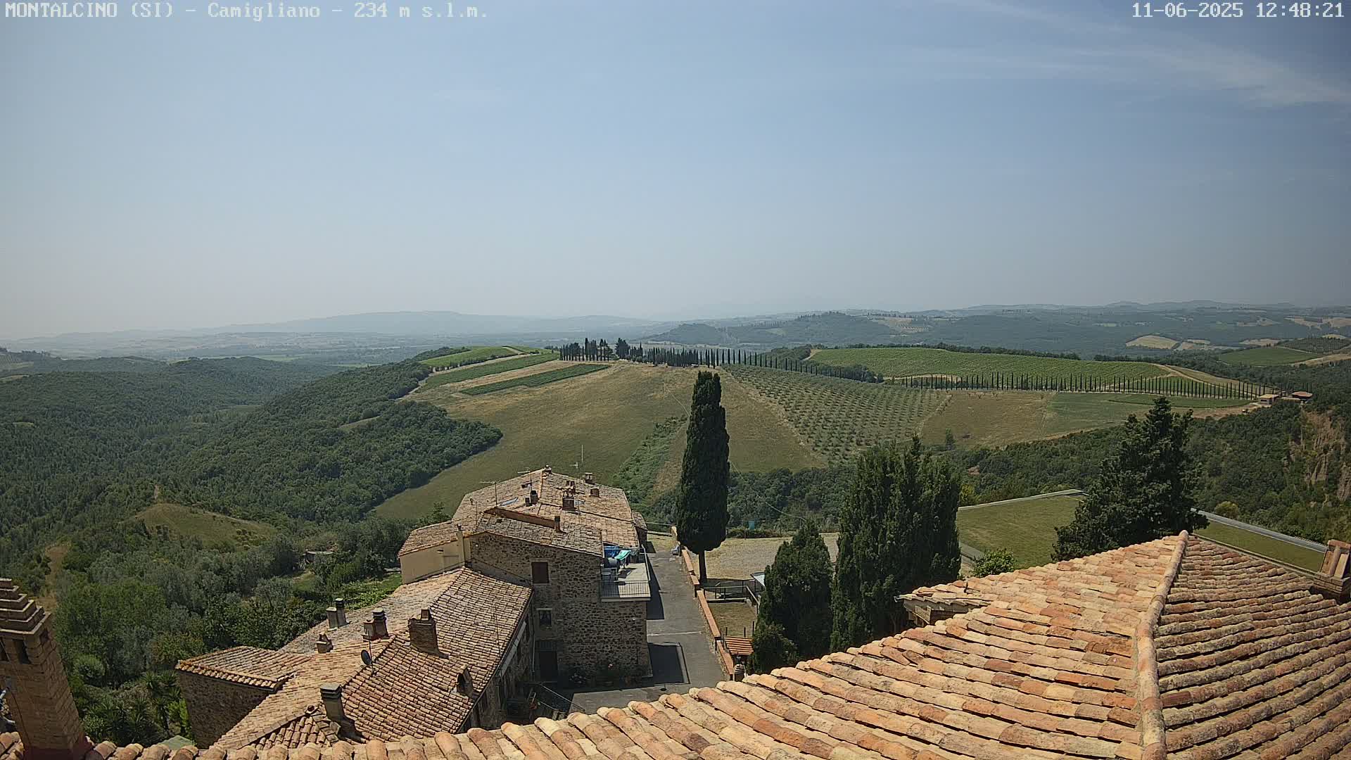 A sunny view from a rooftop shows terracotta-tiled houses overlooking a hilly landscape dotted with cypress trees and vineyards under a clear sky.