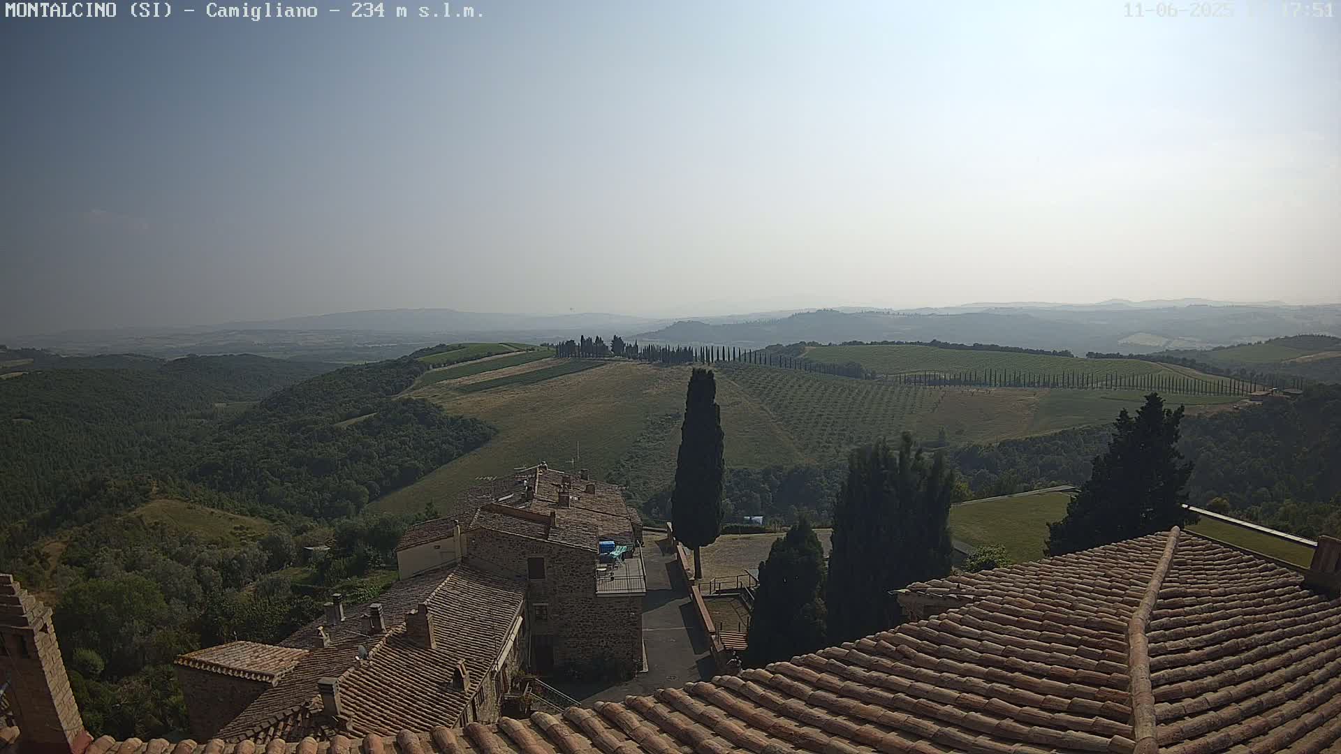 A hazy, sunny view from a terracotta roof shows a Tuscan landscape of rolling hills, olive groves, cypress trees, and stone buildings.