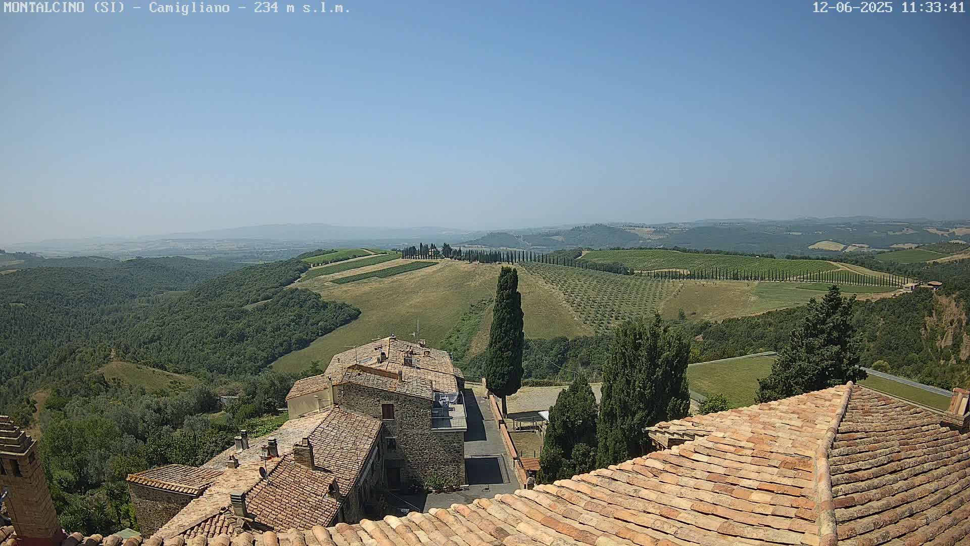 A clear, sunny day reveals a panoramic view of rolling hills dotted with olive groves and vineyards, as seen from a high vantage point overlooking terracotta-roofed buildings.