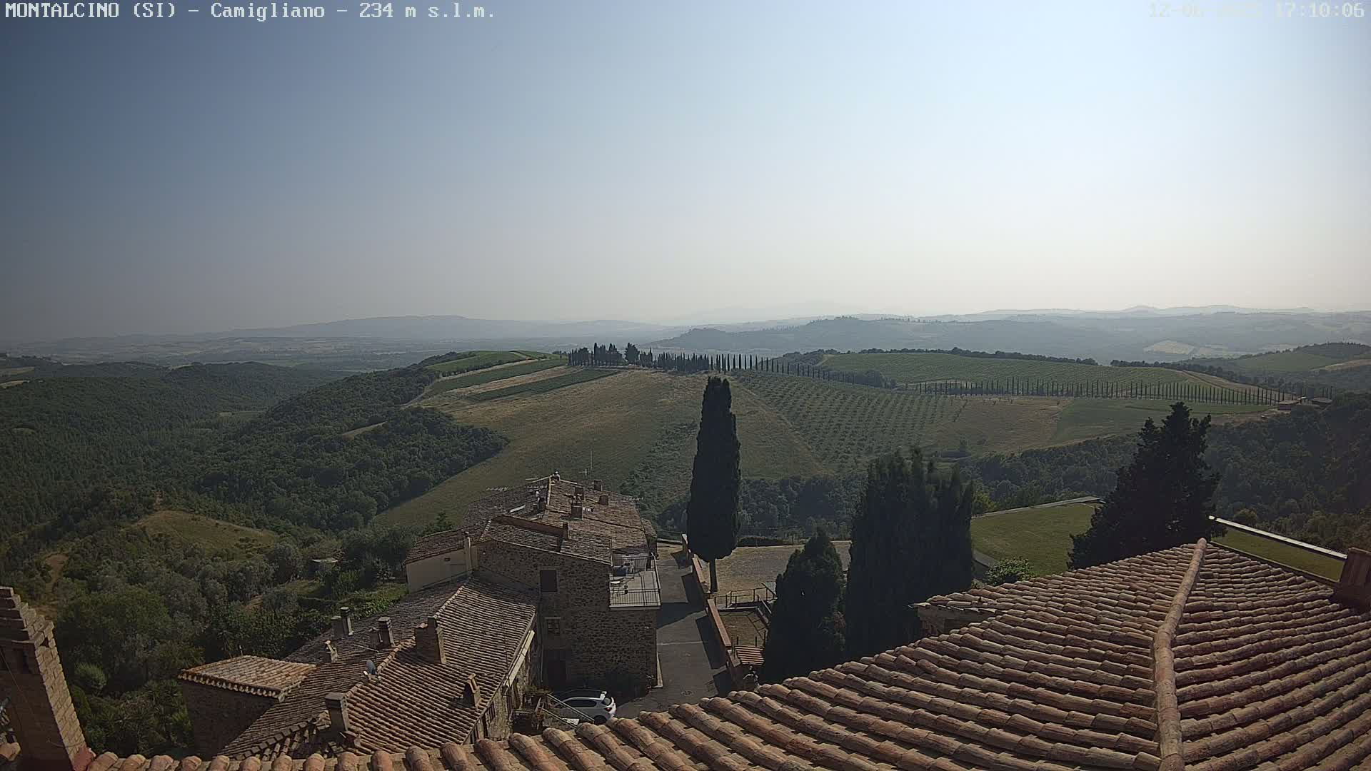 A hazy, sunny day reveals a picturesque Tuscan landscape, featuring rolling hills dotted with olive groves and cypress trees, as seen from a rooftop overlooking a small village with terracotta tile roofs.