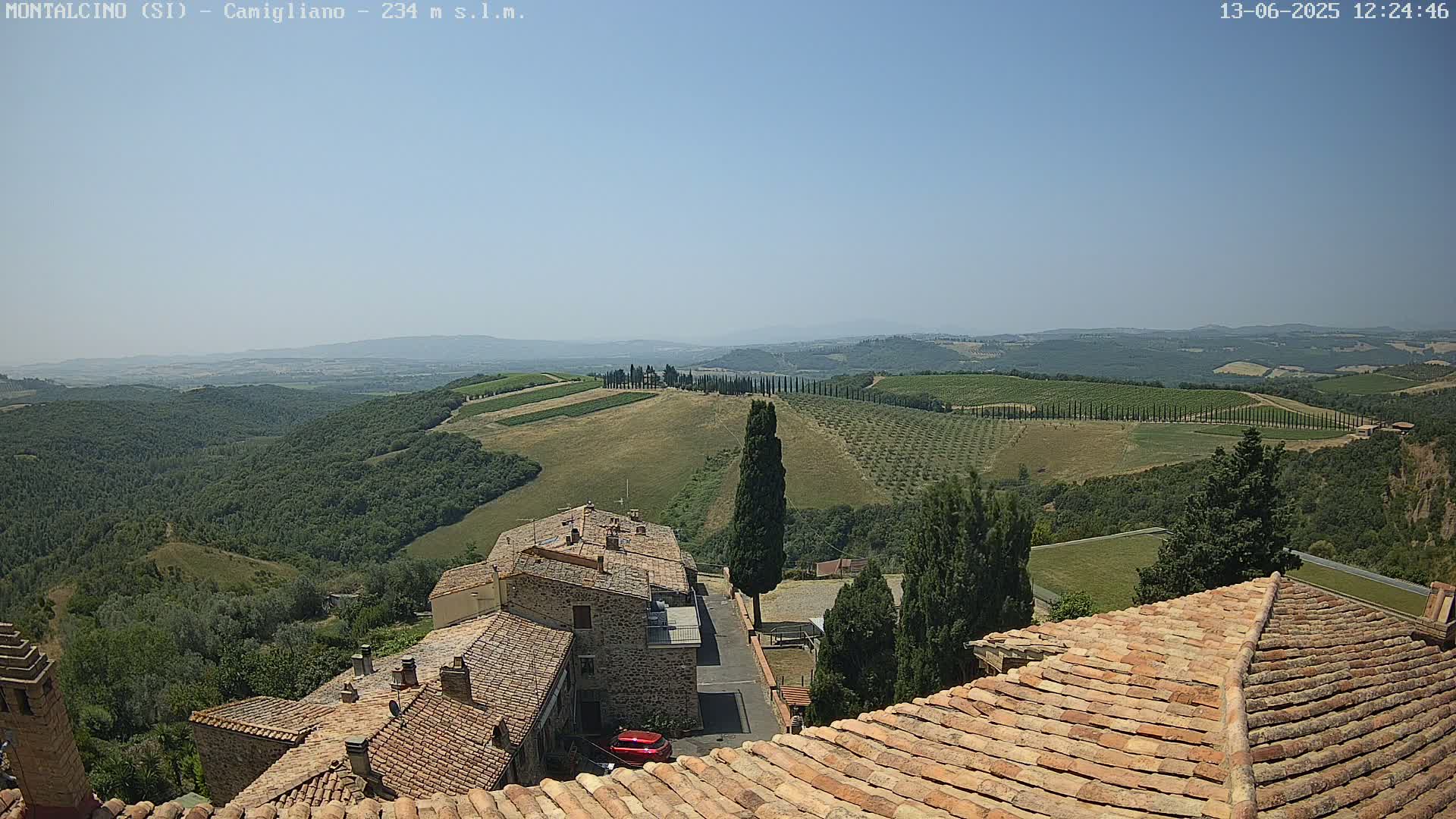 A sunny, clear day reveals a picturesque view of rolling hills dotted with olive groves and cypress trees, overlooking terracotta-roofed buildings in a quaint village.