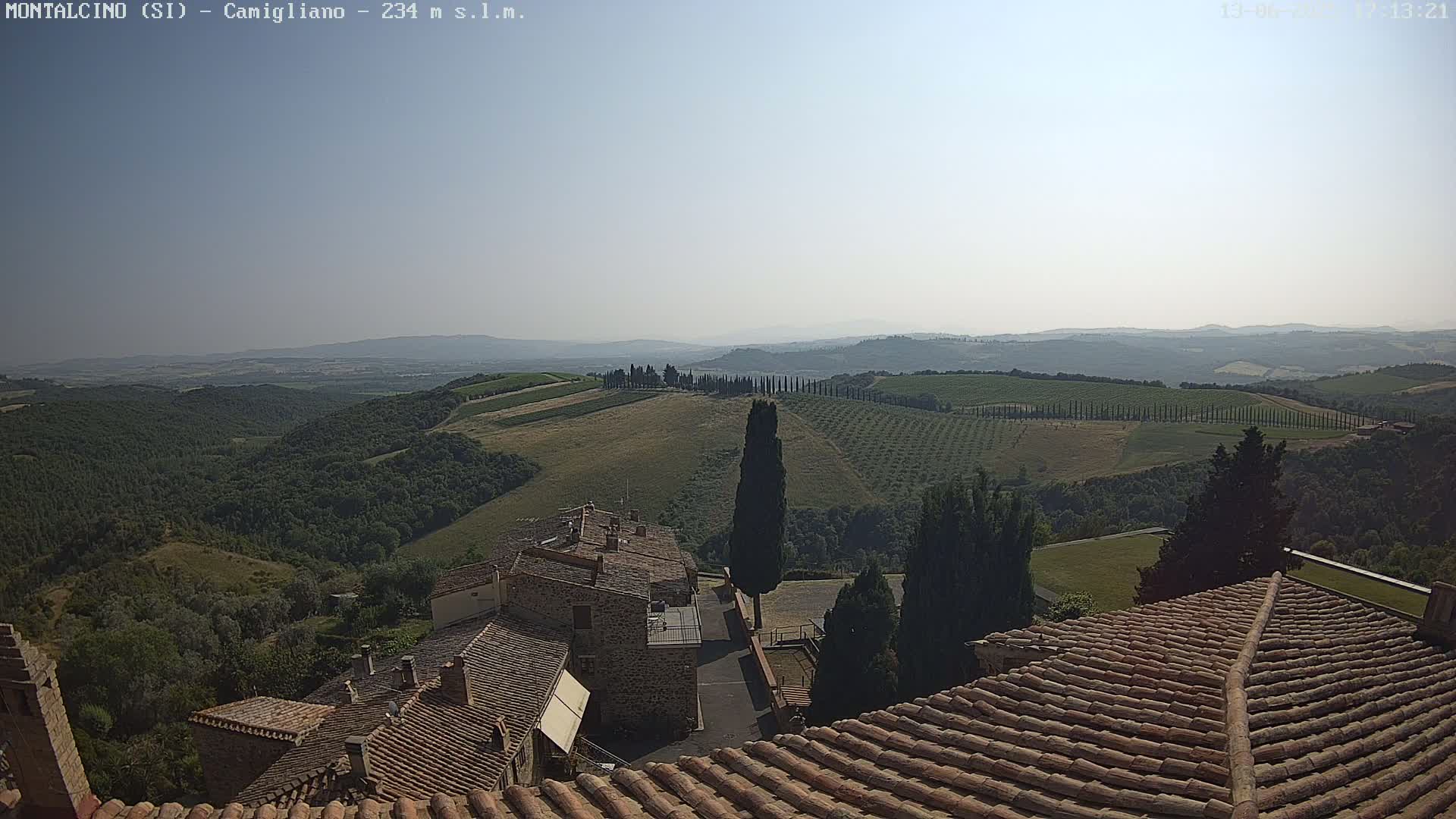 A hazy, sunny day reveals a Tuscan landscape of rolling hills, vineyards, and cypress trees, viewed from a rooftop with terracotta tiles.