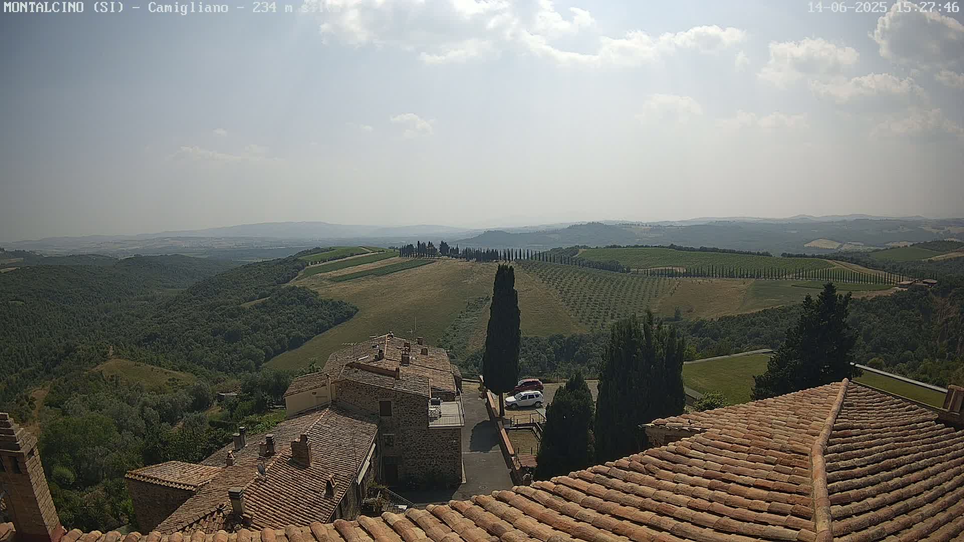 A partly cloudy, sunny day overlooks terracotta-roofed buildings nestled in a hilly landscape with vineyards and rows of cypress trees.