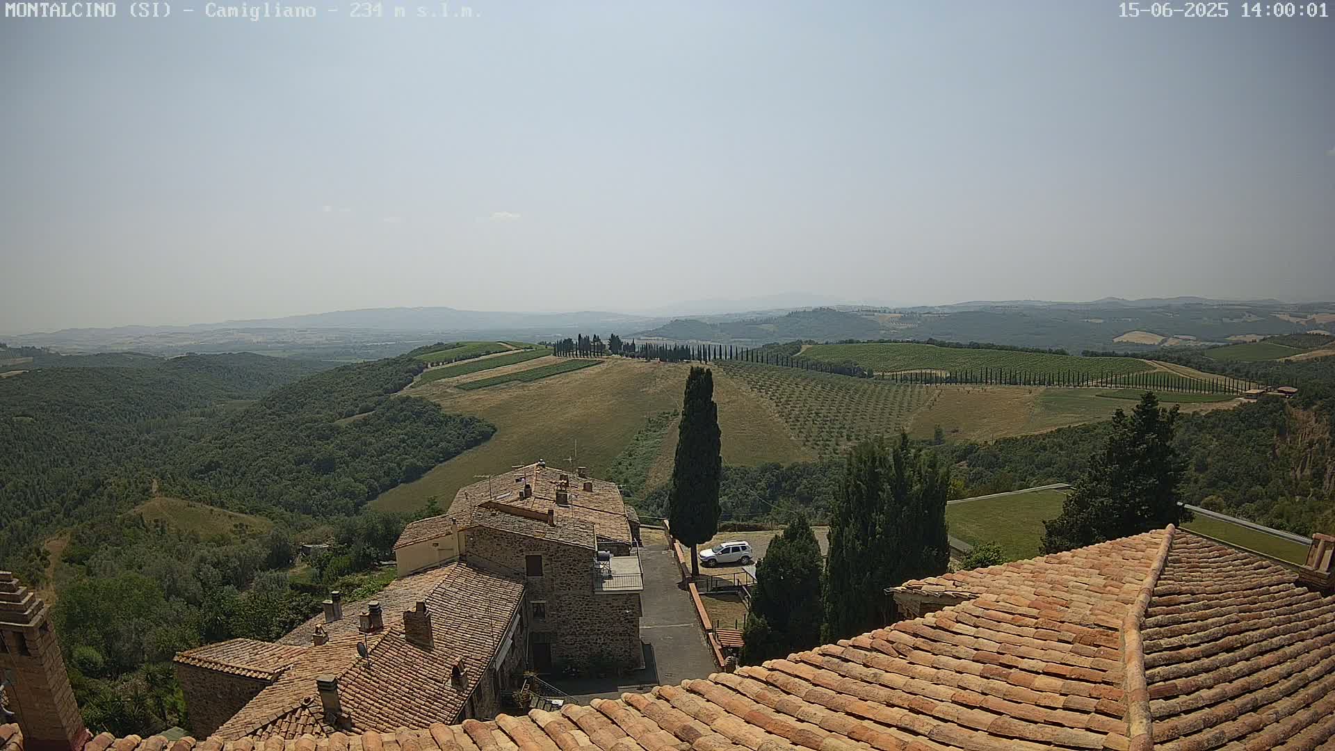 A view from a terracotta roof shows stone buildings and a sunny, hazy landscape of rolling hills, vineyards, and olive groves.