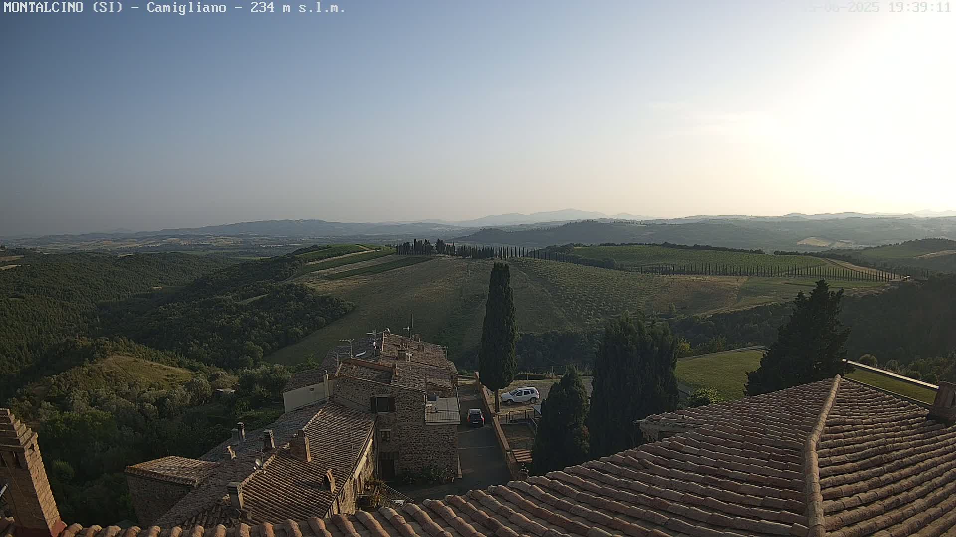 A view from a terracotta roof shows a hazy, sunny day over rolling hills dotted with trees and buildings in a rural landscape.