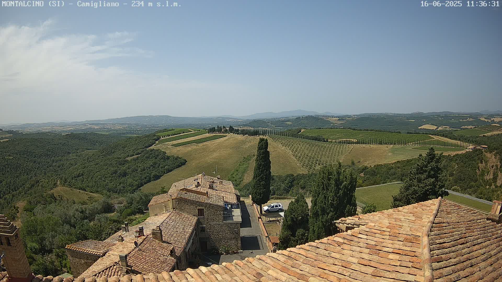 A sun-drenched view from a tiled rooftop shows a Tuscan landscape of rolling hills, vineyards, and olive groves, with a small village nestled below.