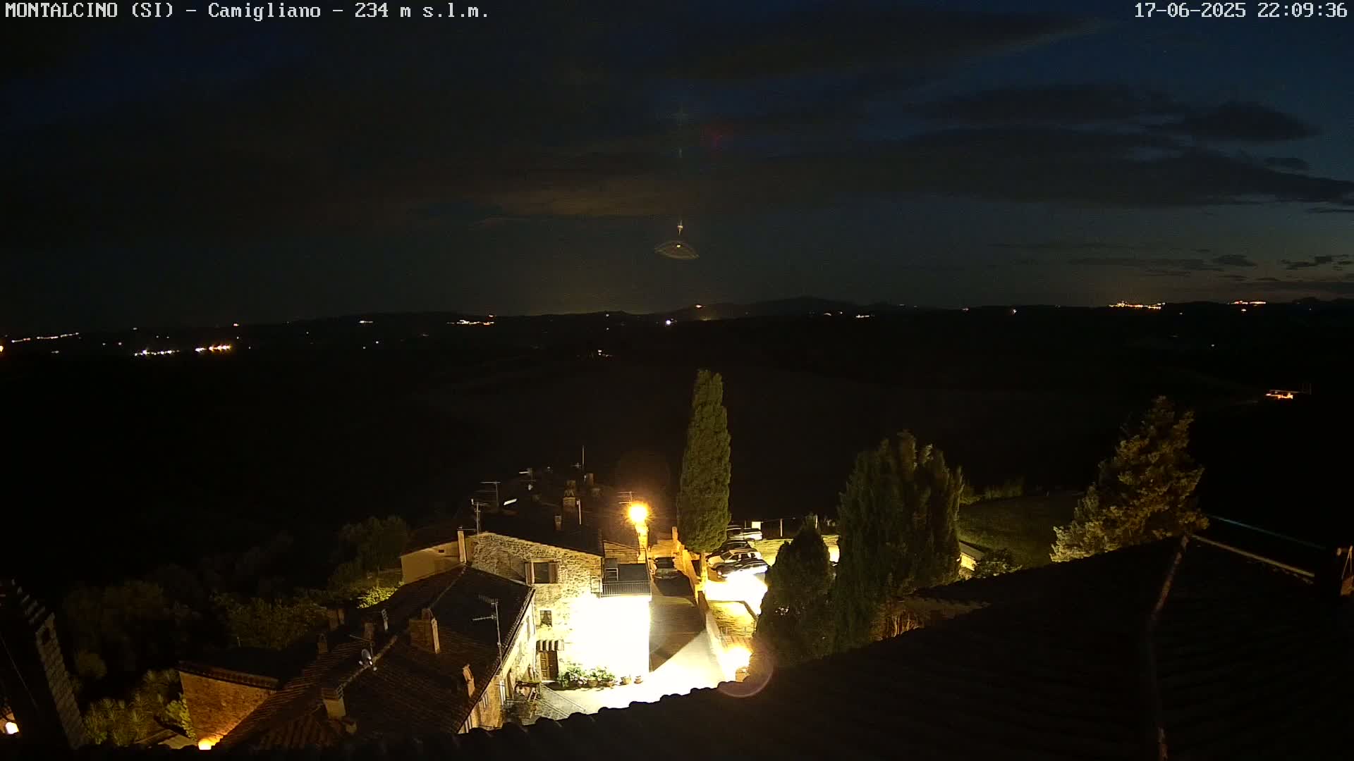 An illuminated village at night, under a mostly clear, dark sky, overlooks a distant, dimly lit landscape.