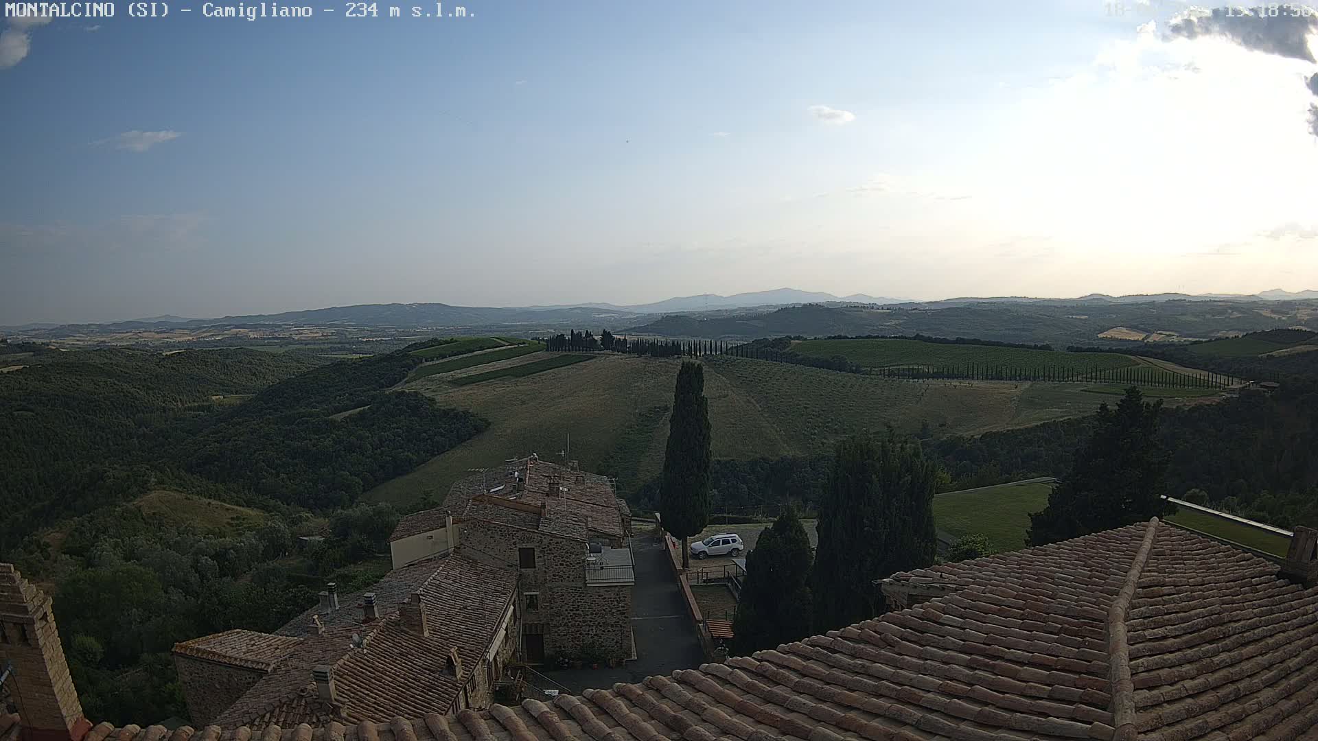 Under a mostly clear sky, a view from a rooftop shows rolling hills of vineyards and olive groves, and a small village with terracotta roofs.