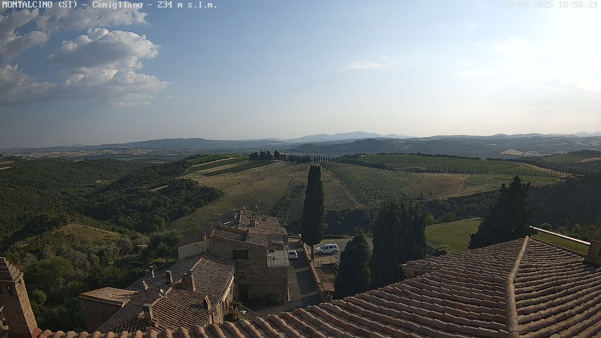 A partly cloudy sky overlooks a hilly landscape of vineyards and olive groves, with a village of terracotta-roofed buildings in the foreground.