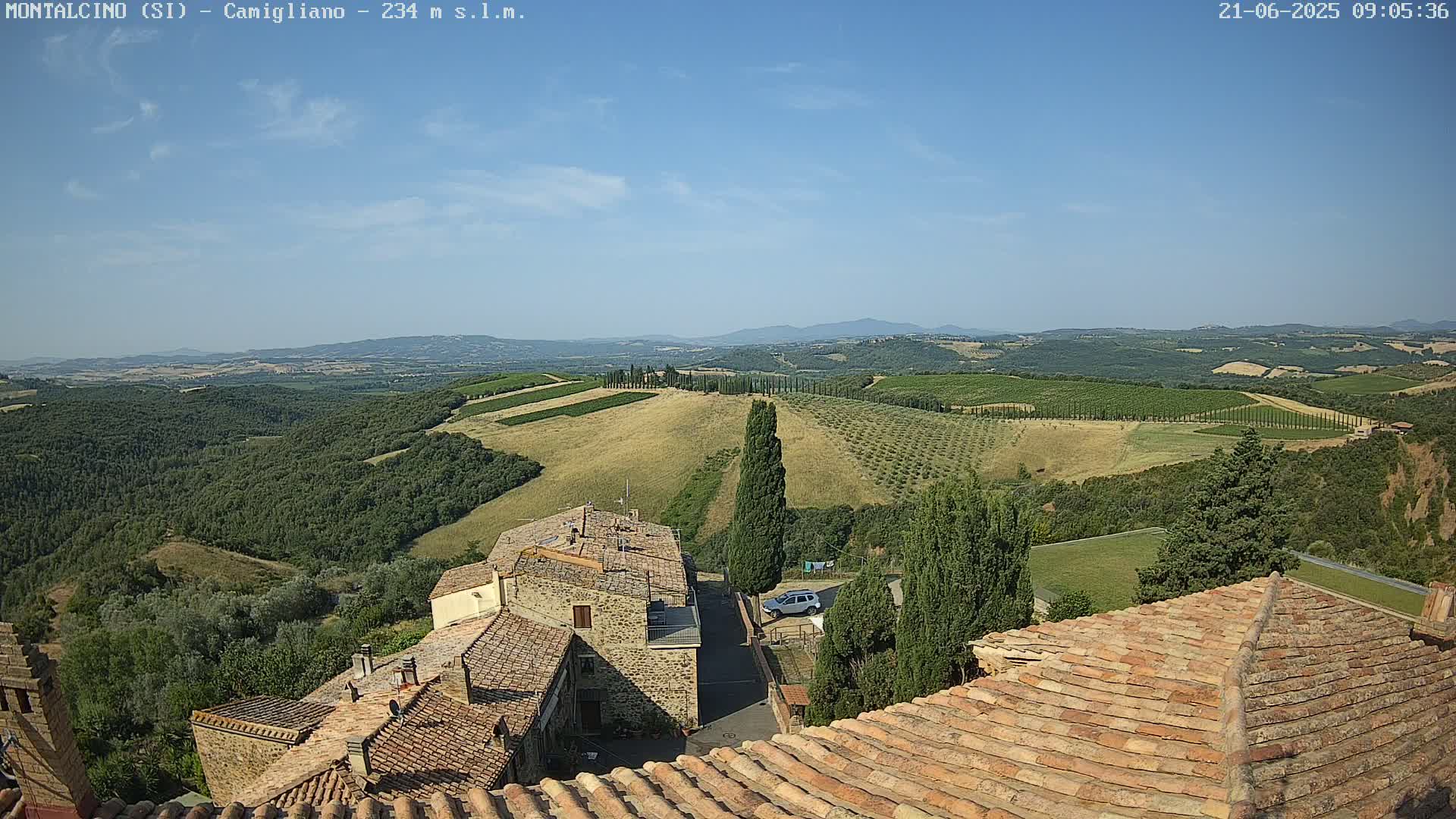 A clear, sunny day reveals a panoramic view of rolling hills dotted with vineyards and olive groves, overlooking a village with terracotta roofs.