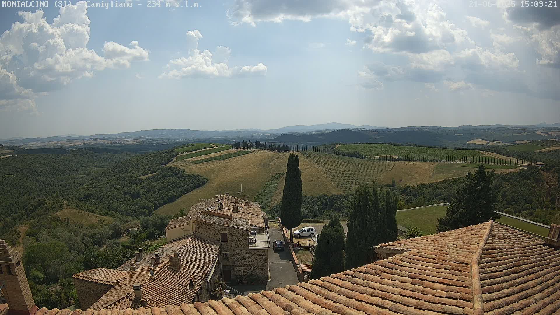 A partly cloudy day reveals a view of rolling hills, vineyards, and a stone village, seen from a rooftop.