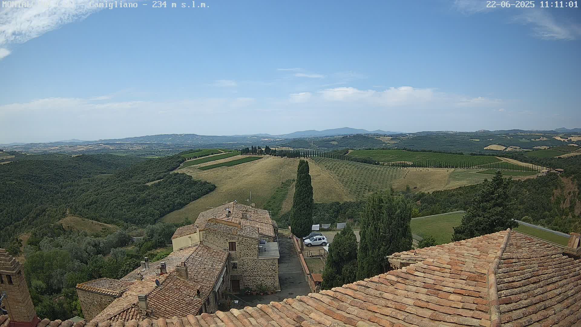 A partly cloudy blue sky overlooks a hilly landscape of vineyards and stone buildings with red-tiled roofs.