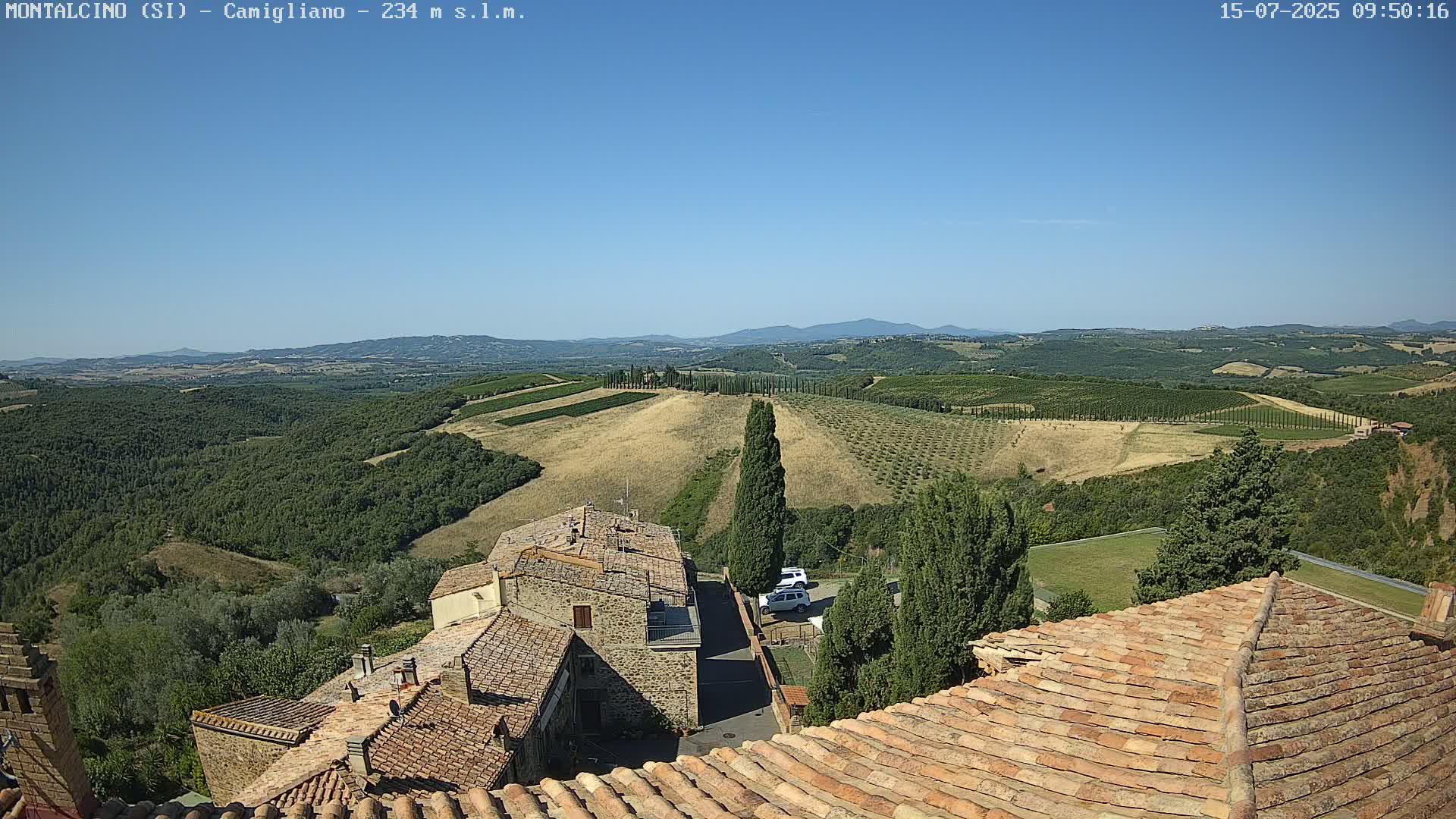 A clear, sunny day reveals a Tuscan landscape of rolling hills dotted with olive groves and cypress trees, viewed from a rooftop overlooking stone buildings with terracotta tile roofs.