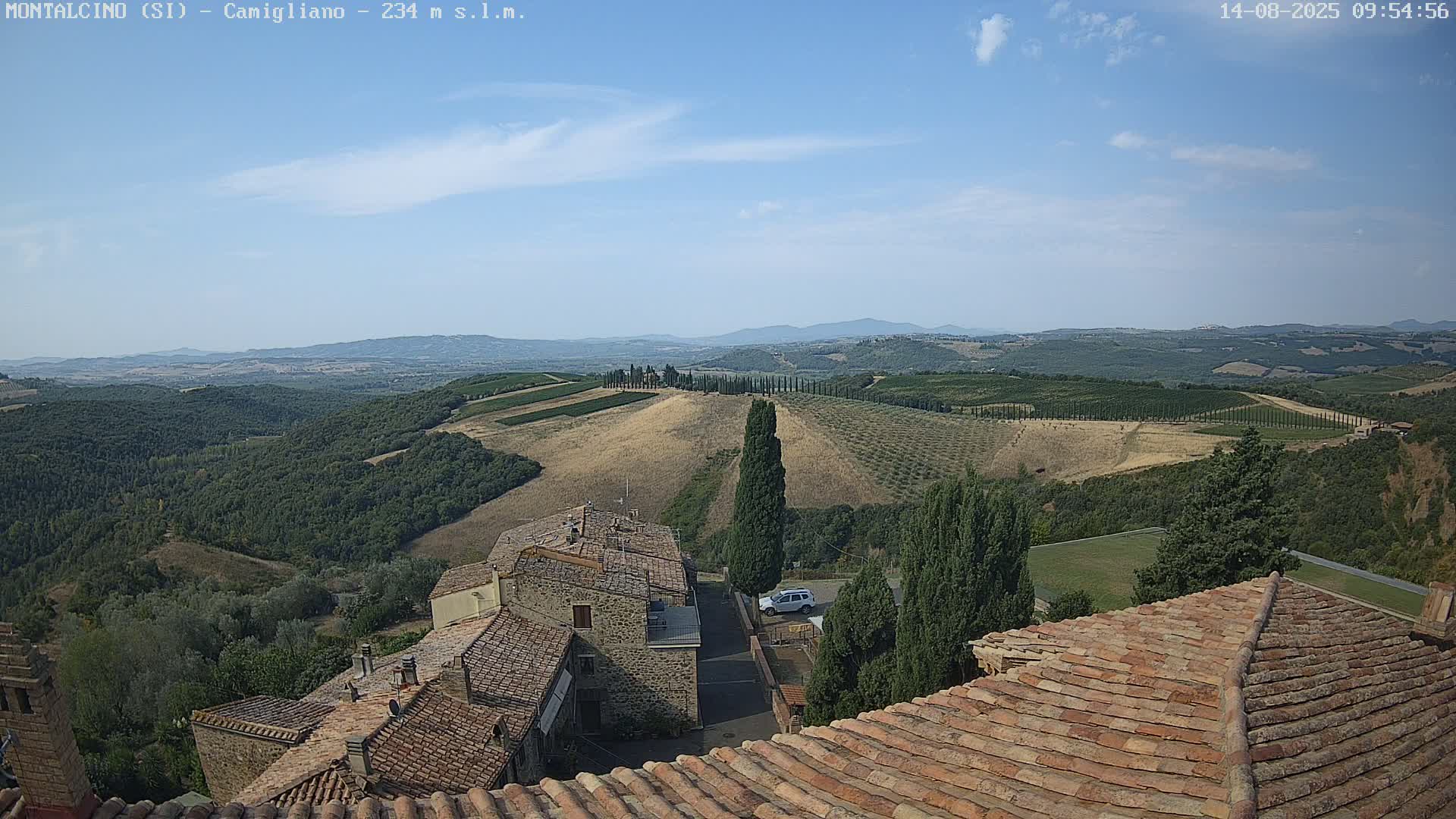 A view from a rooftop shows terracotta roofs of buildings, a small village, and a sunny, expansive landscape of rolling hills, vineyards, and olive groves under a mostly clear blue sky.