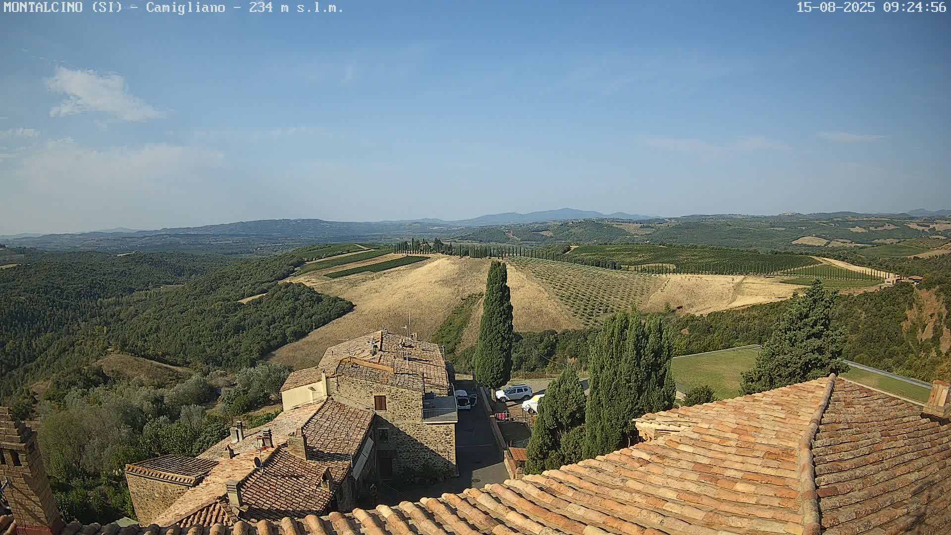 A high-angle view on a clear day shows terracotta rooftops of buildings in a village overlooking rolling hills with vineyards and cypress trees under a mostly sunny sky.