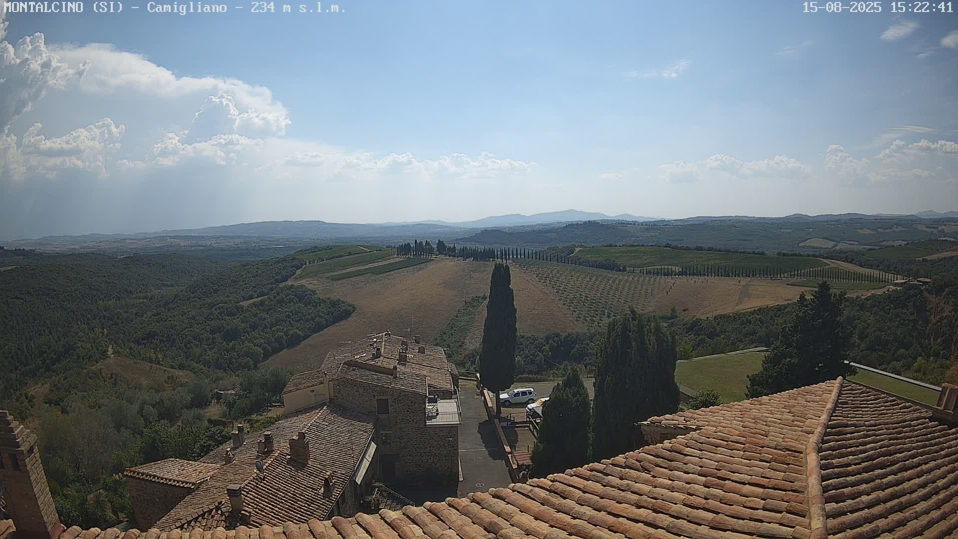 A high-angle view on a clear day shows terracotta rooftops of buildings in a village overlooking rolling hills with vineyards and cypress trees under a mostly sunny sky.