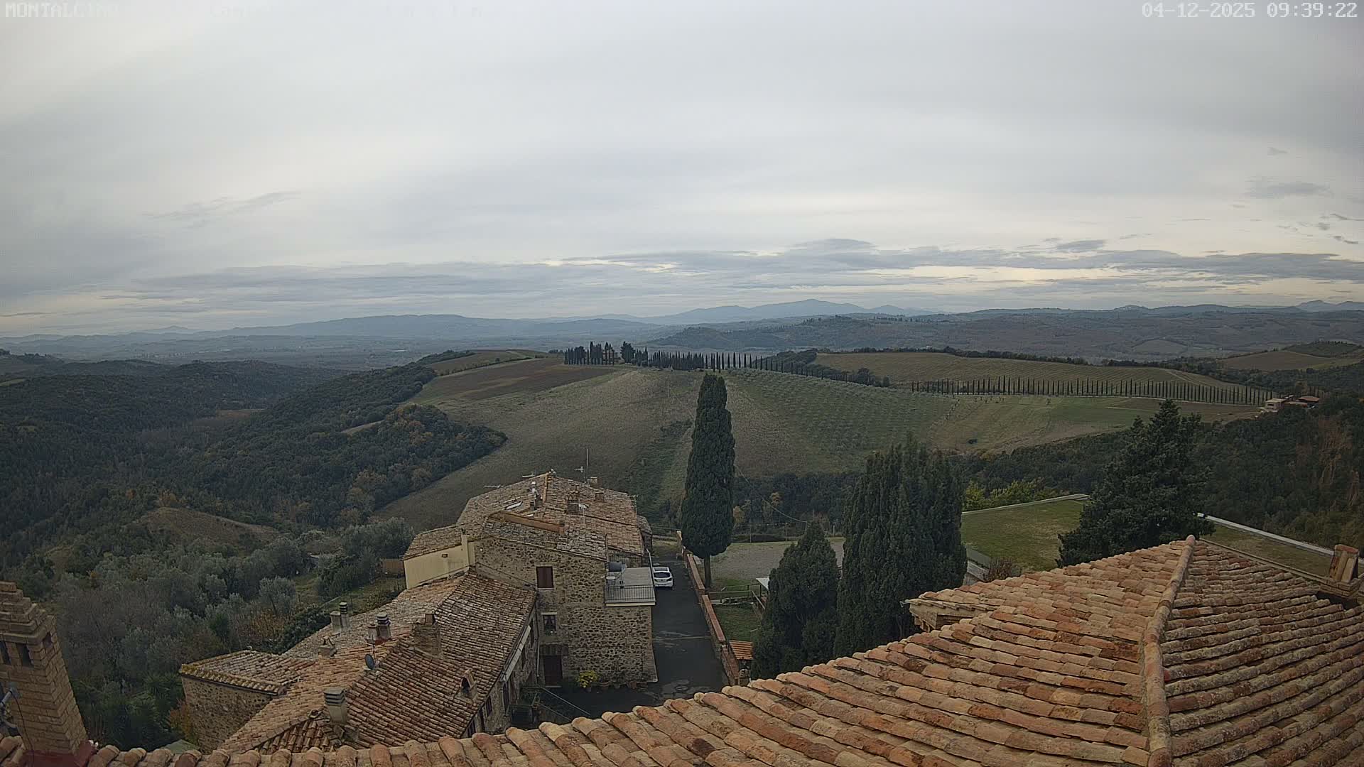 Terracotta-tiled rooftops in the foreground overlook a vast, rolling Tuscan landscape of fields, forests, and distinct rows of cypress trees, all under a uniformly overcast and cloudy sky with distant hills fading into the horizon.