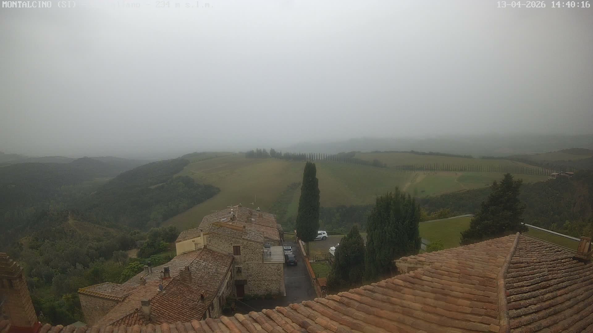 Old tiled rooftops and tall cypress trees offer a view over sunlit rolling hills, cultivated fields, and rows of trees, all rising above a dense valley fog under a clear blue sky.